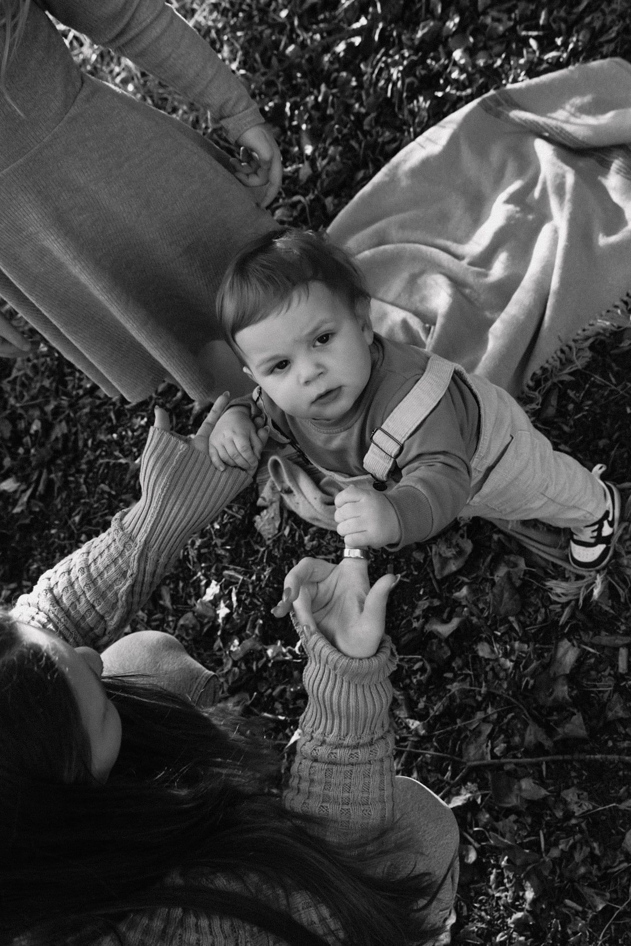 mom helping toddler stand and holding his hands as he looks up at camera