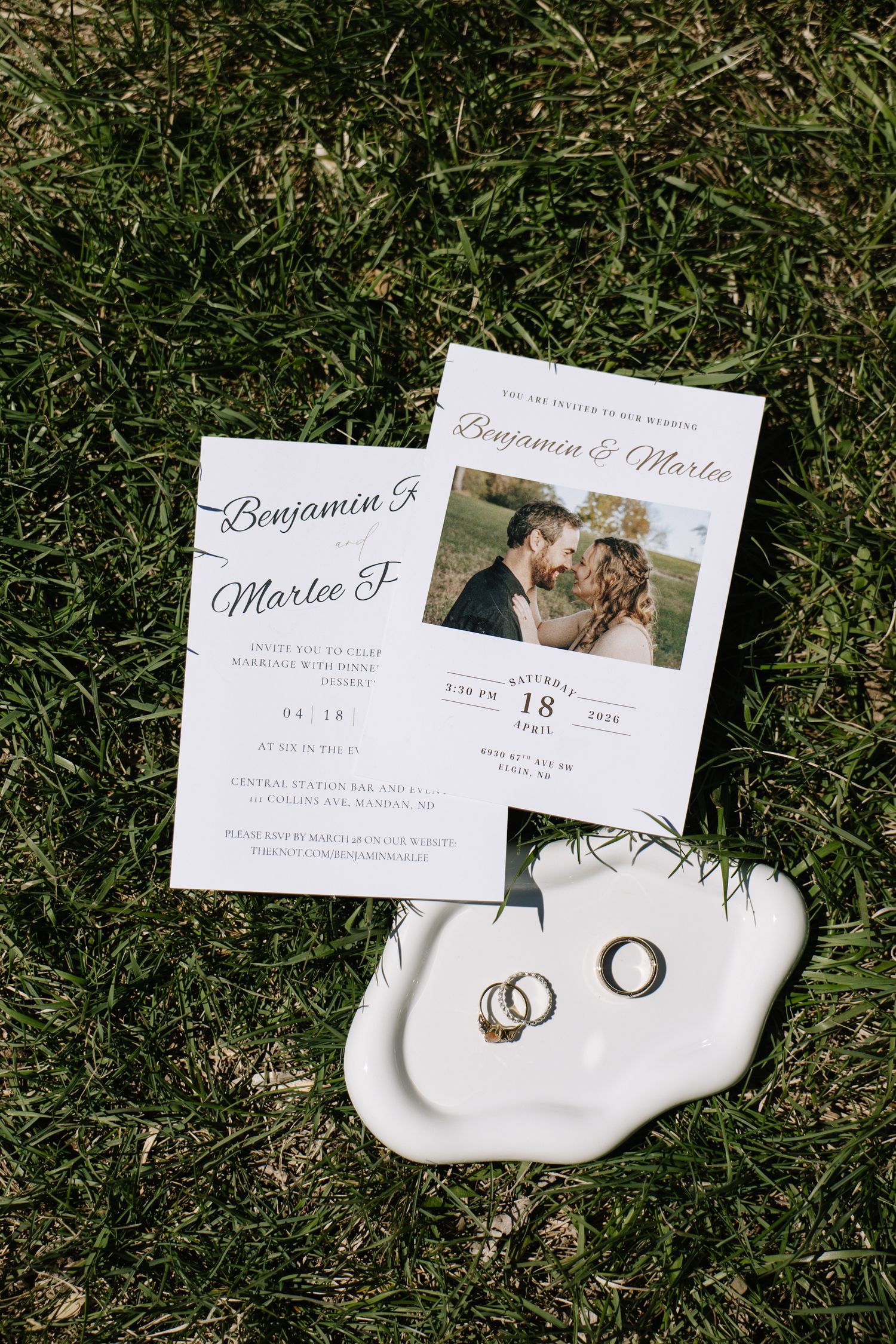 Wedding invitations for Benjamin and Marlee displayed on green grass alongside gold and silver wedding rings on a white dish.
