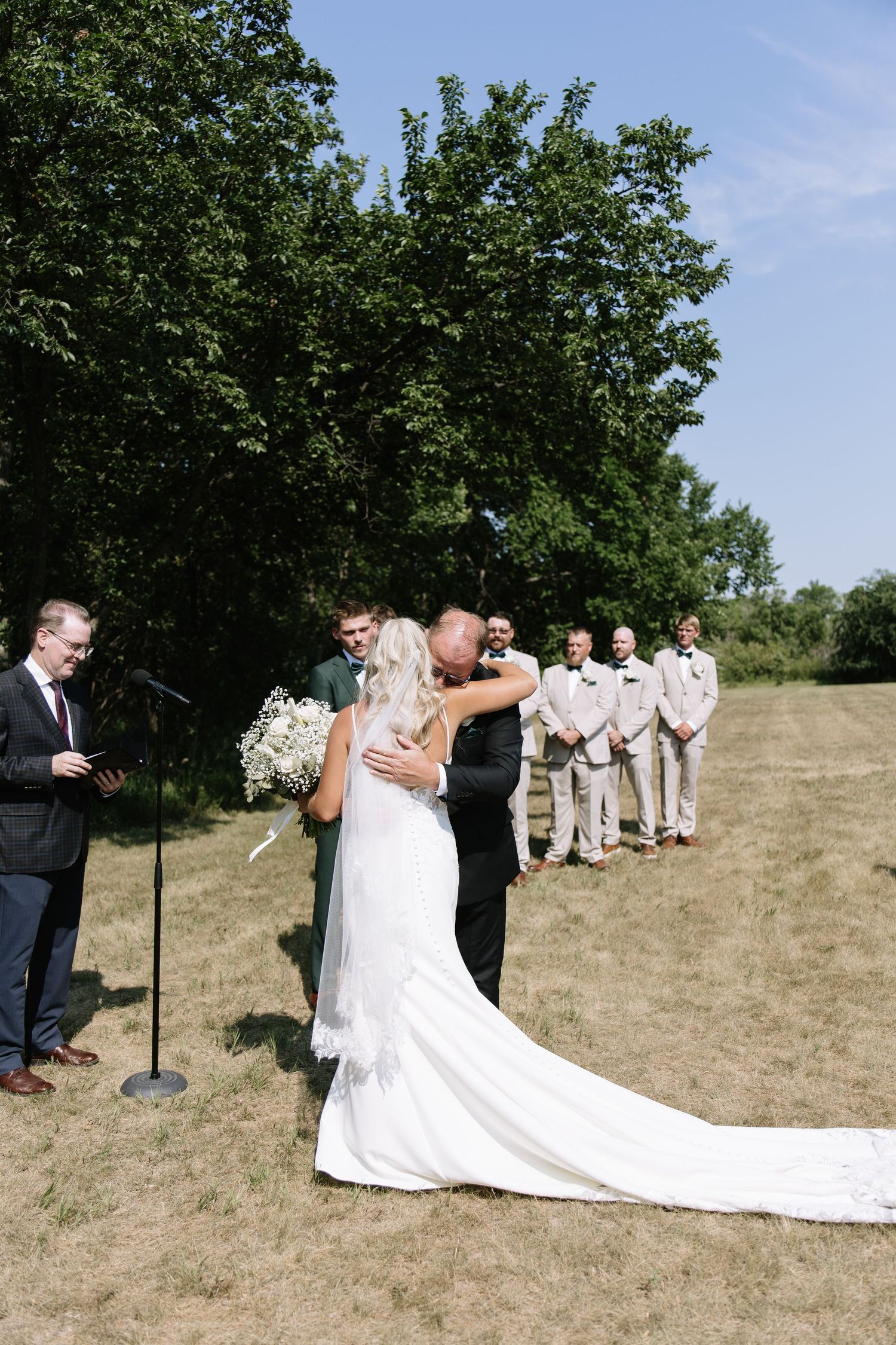 Bride and groom share first kiss at outdoor wedding ceremony with groomsmen and officiant on sunny day.