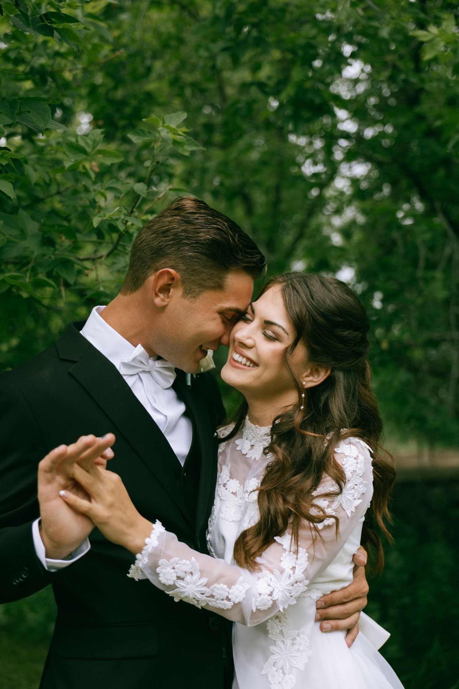 A smiling bride in a lace gown and groom in a black tuxedo share a romantic dance outdoors among lush green trees.