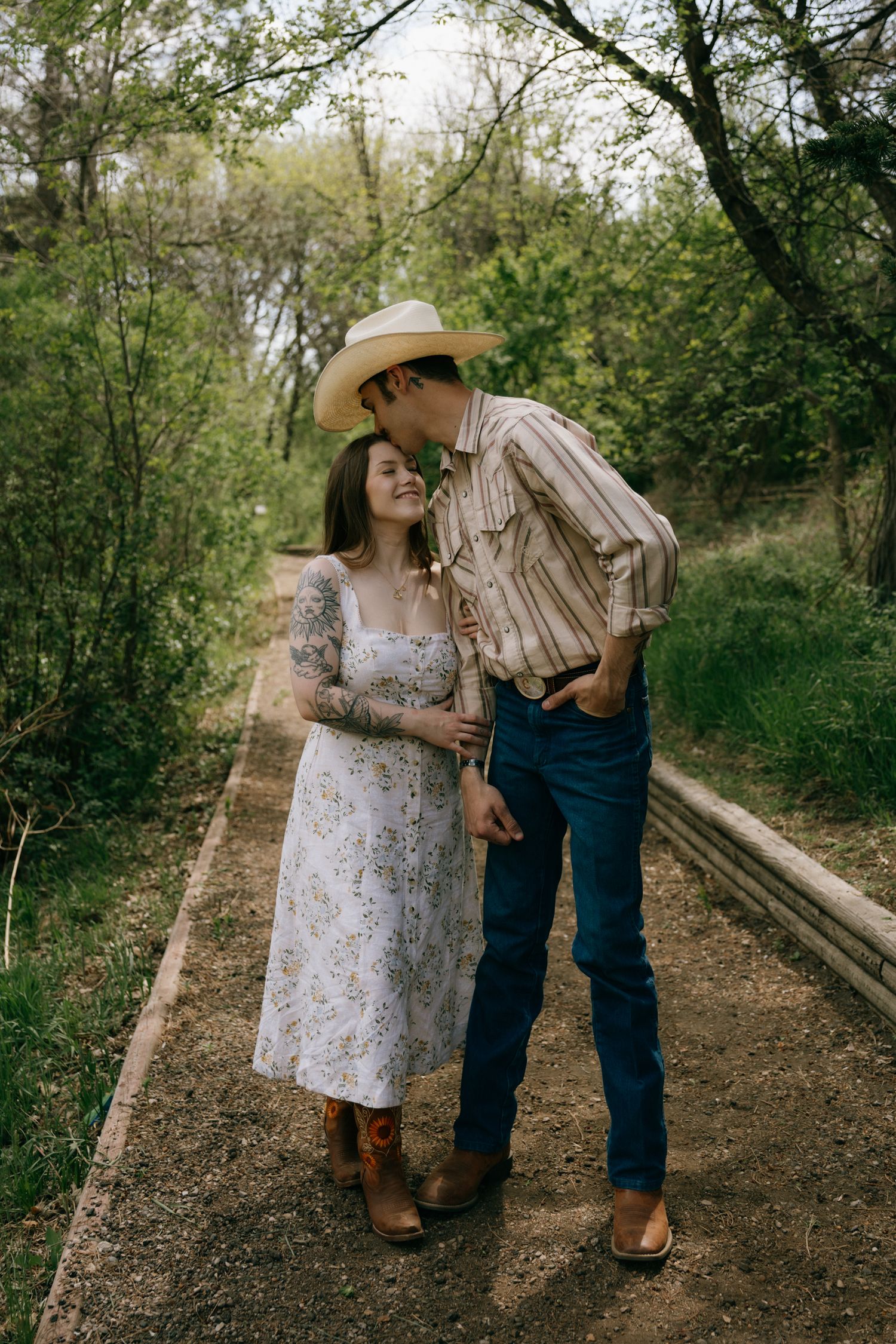 A couple in western attire share a tender moment on a wooded path, woman in floral dress, man in cowboy hat.