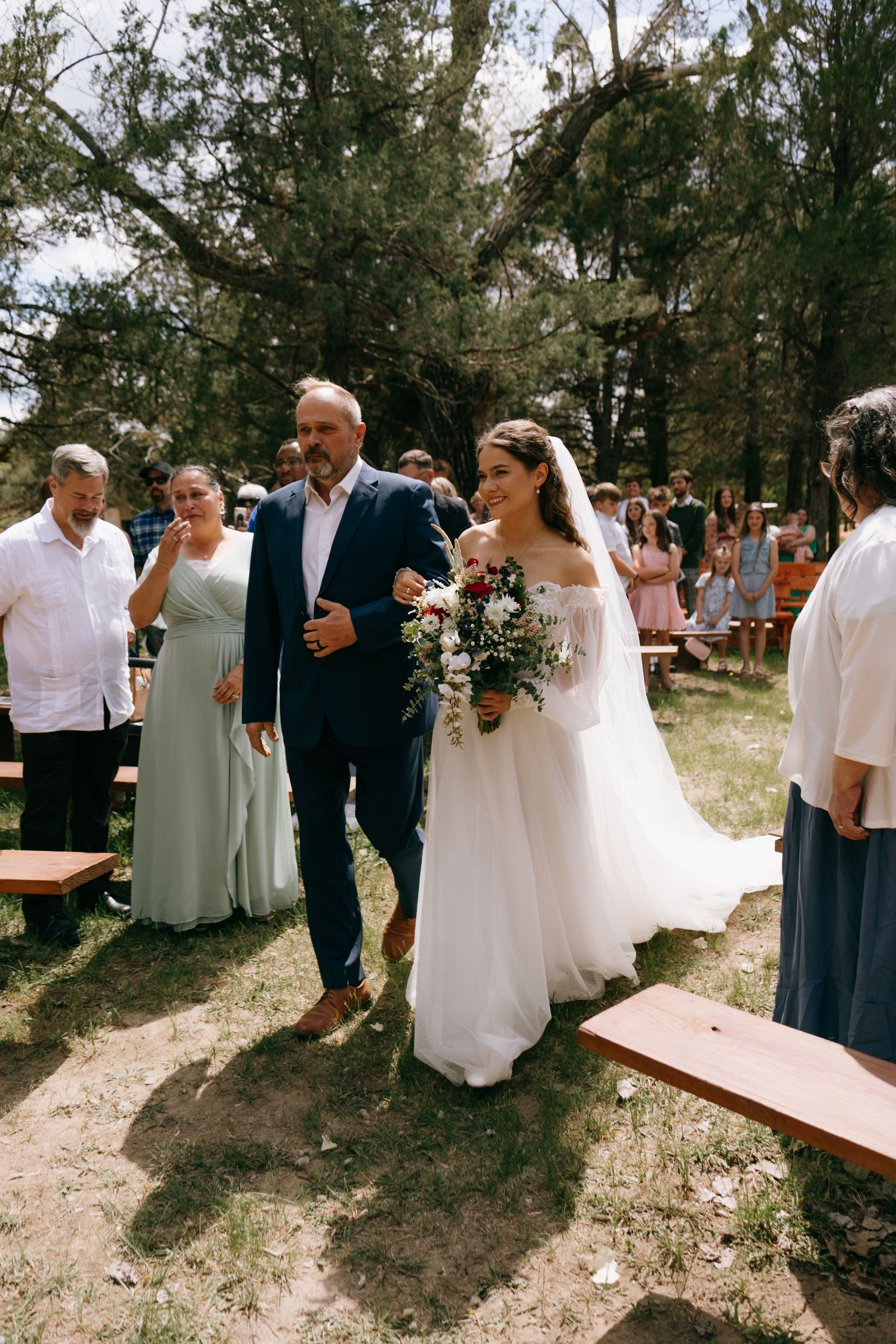 Bride in white gown walks down outdoor aisle with escort, holding bouquet, guests seated among trees.