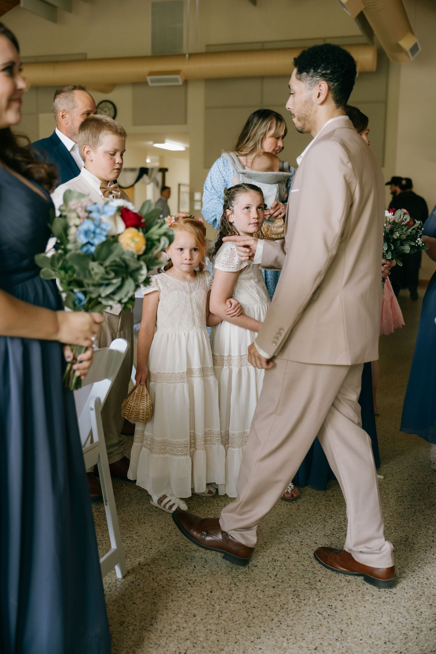 Groom in beige suit walks with flower girls in white dresses during wedding ceremony procession indoors.