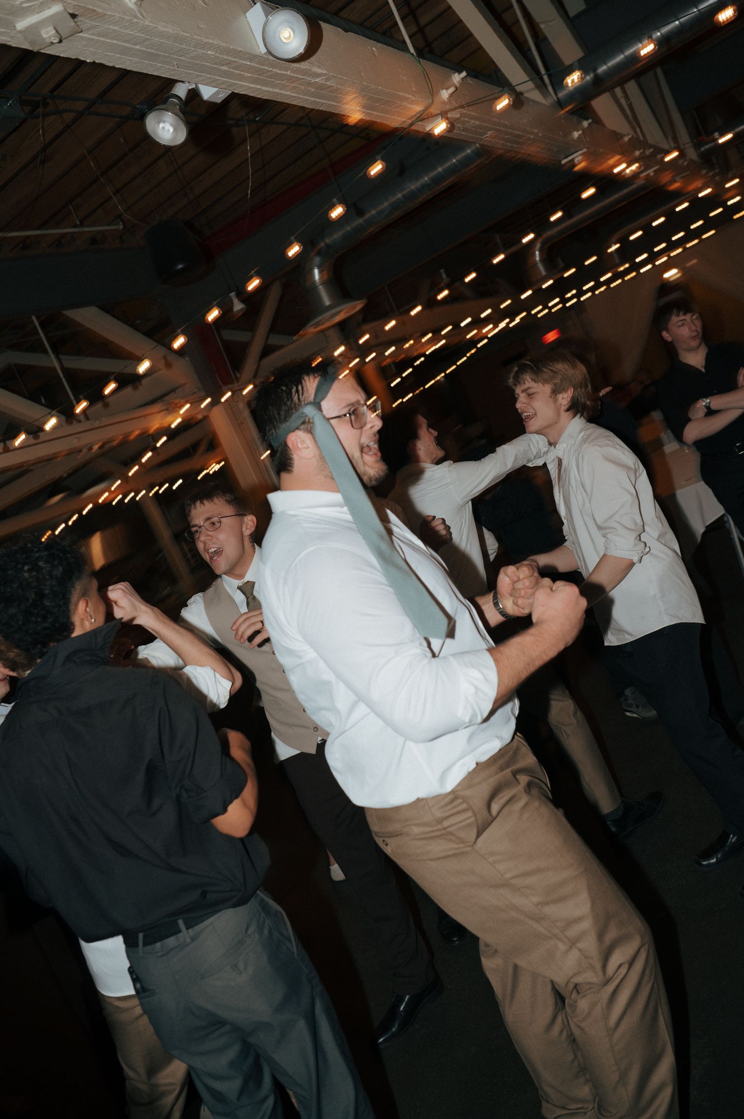 People dancing at an indoor event venue decorated with string lights, wearing formal attire including white shirts and ties.