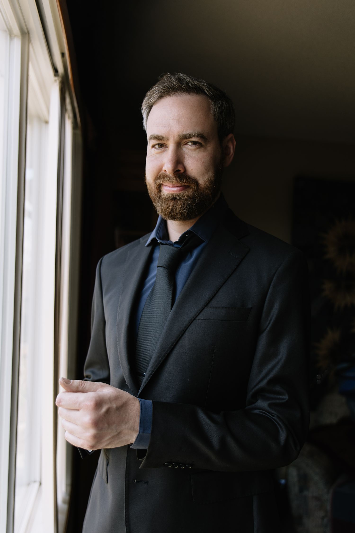 A bearded man in a black suit and tie stands confidently by a window in a dimly lit room.