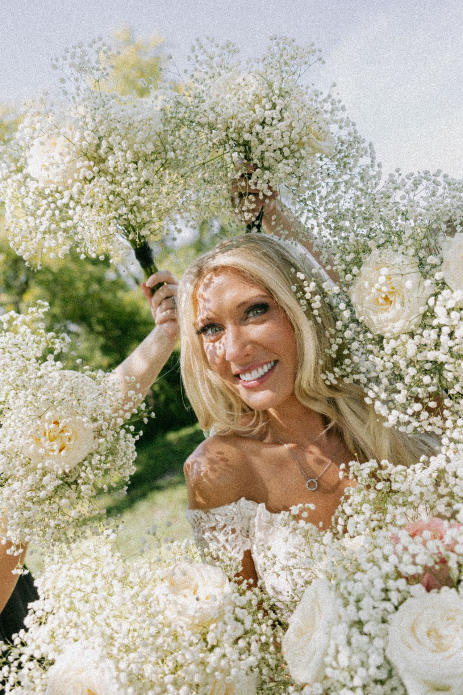 Smiling blonde bride surrounded by white baby's breath and rose bouquets outdoors on her wedding day.