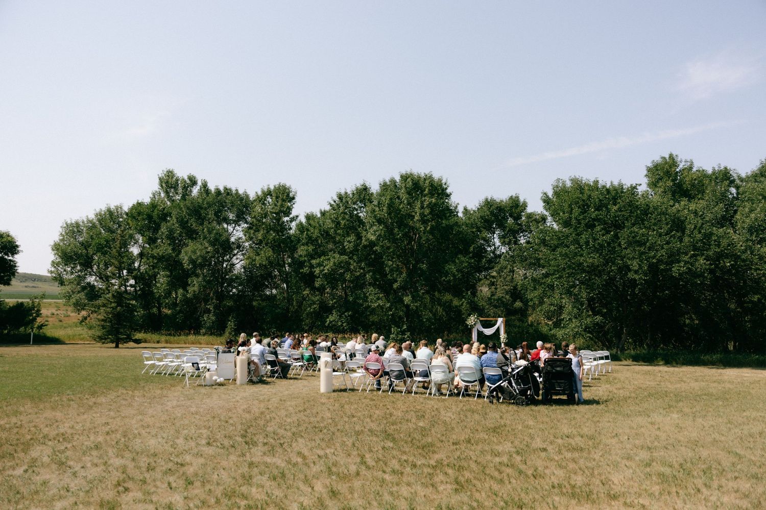 Outdoor wedding ceremony in a green field surrounded by tall trees on a sunny day.