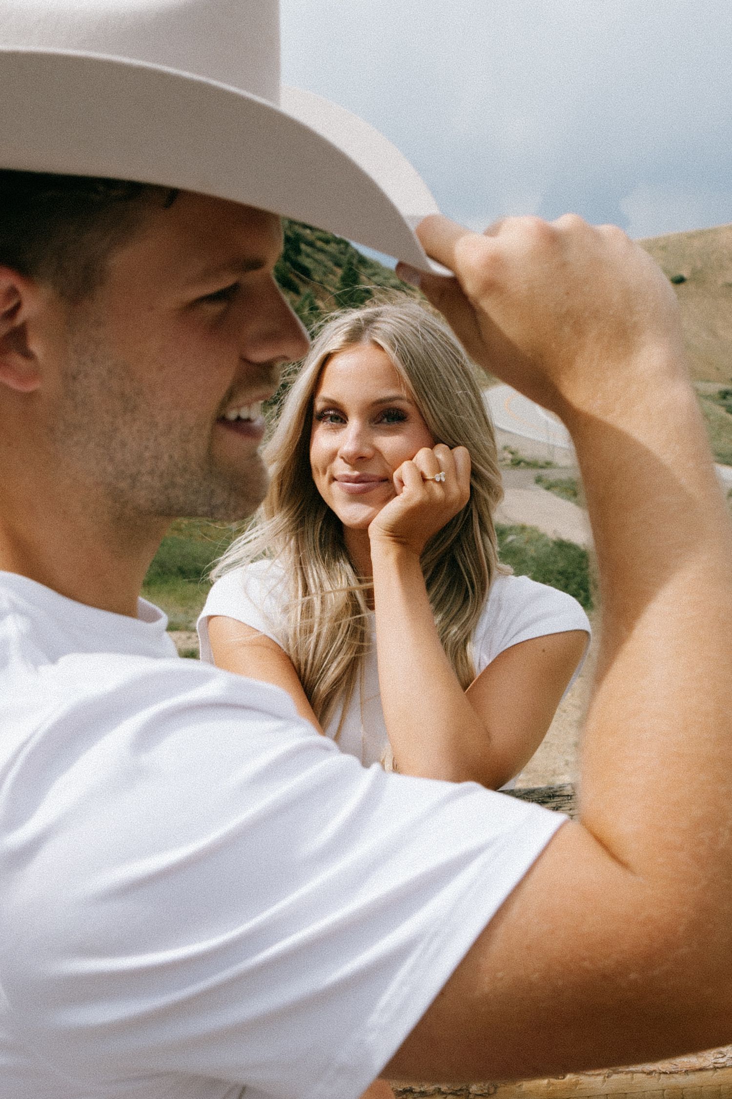 A smiling couple in white t-shirts outdoors, man in cowboy hat tilting brim while woman rests chin on hand gazing at him.