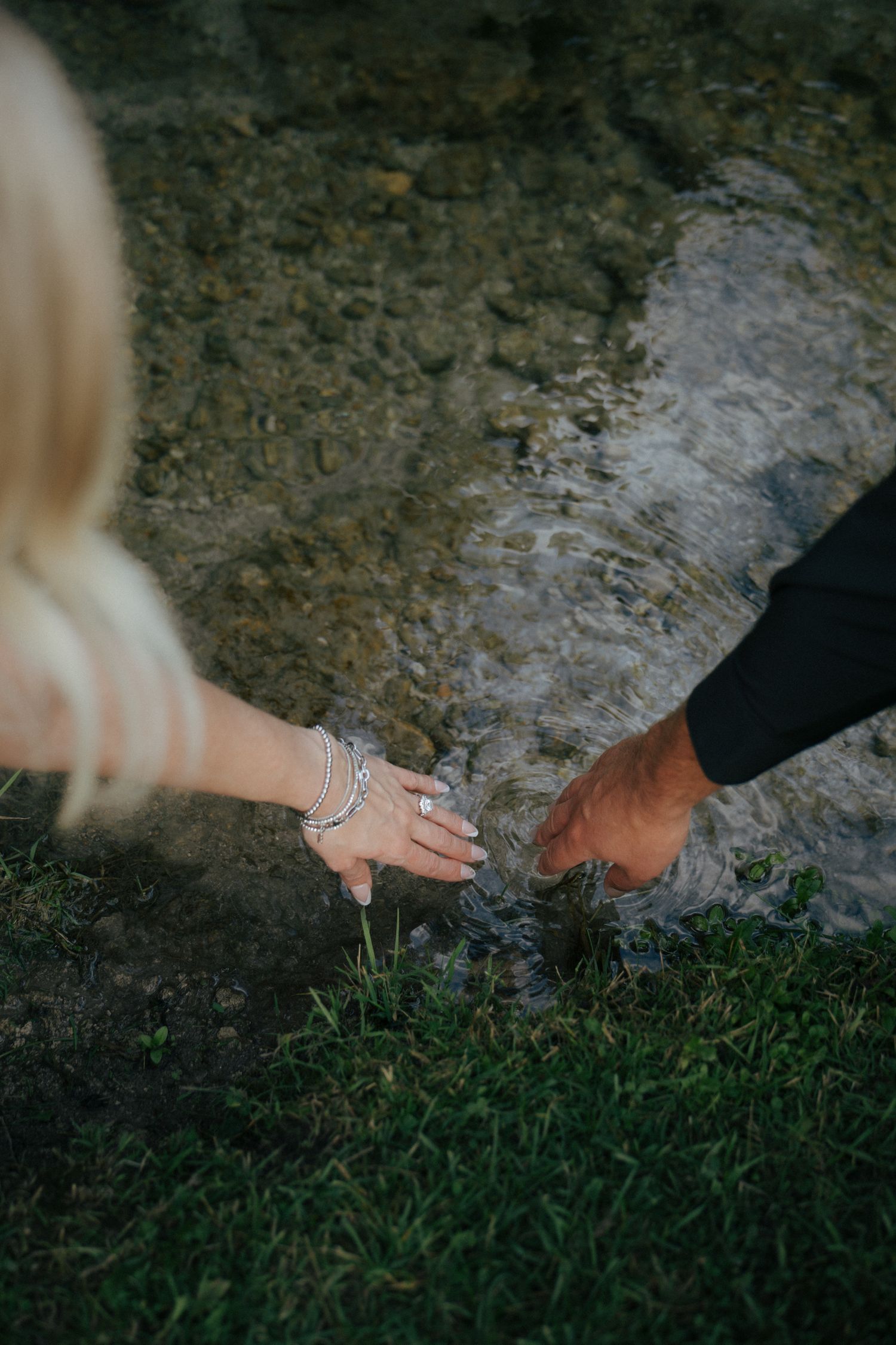 Close-up of hands with engagement ring touching on grass and stone surface.