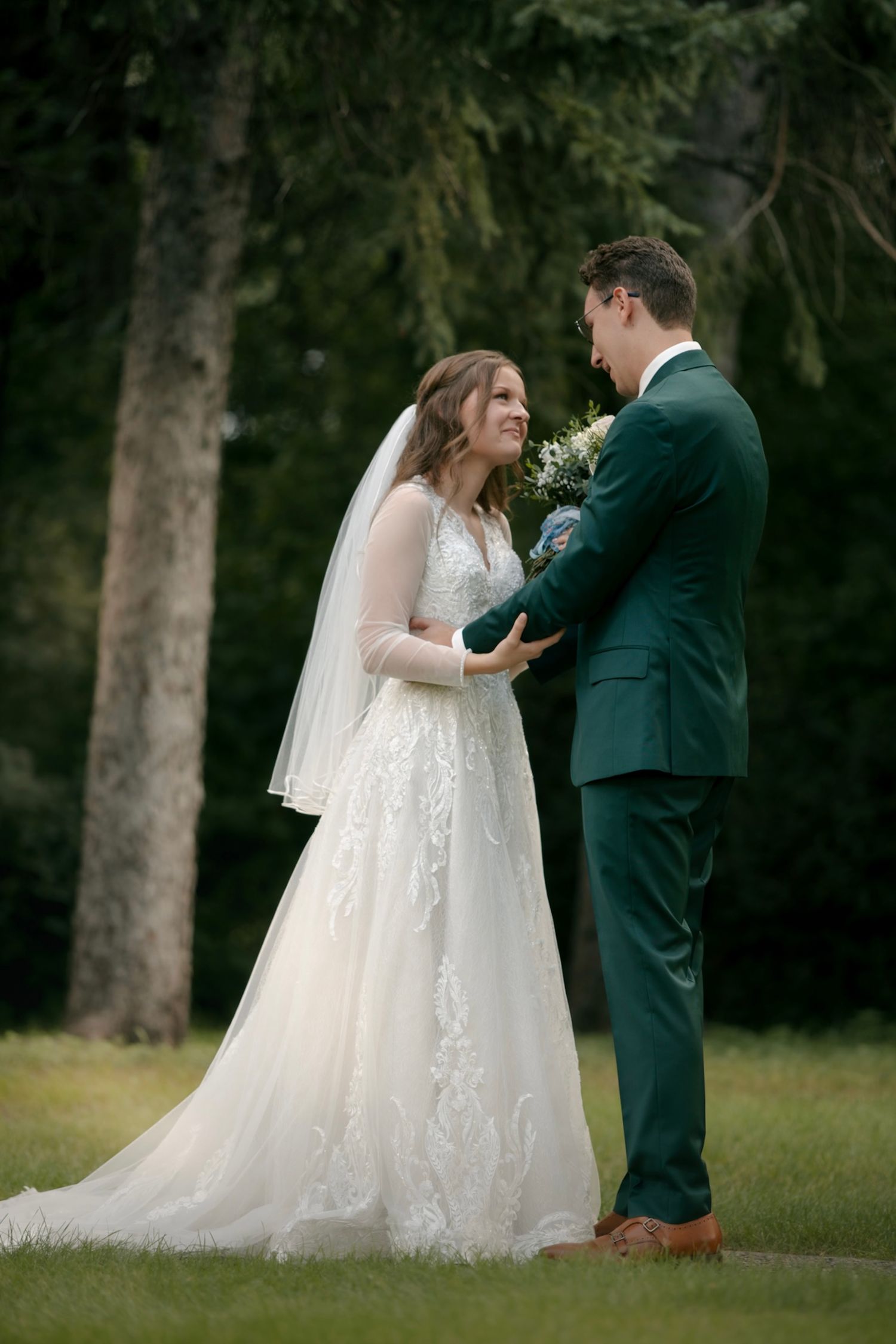 Wedding couple in elegant attire sharing a tender moment outdoors among trees.