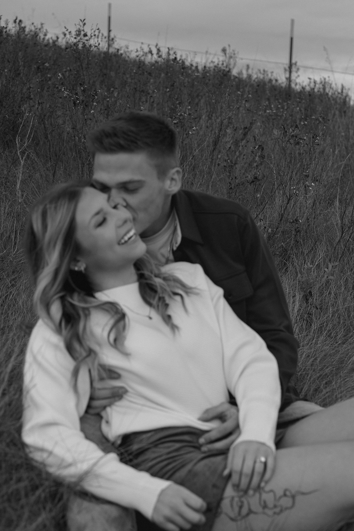 Black and white photo of couple lying in tall grass sharing intimate moment.