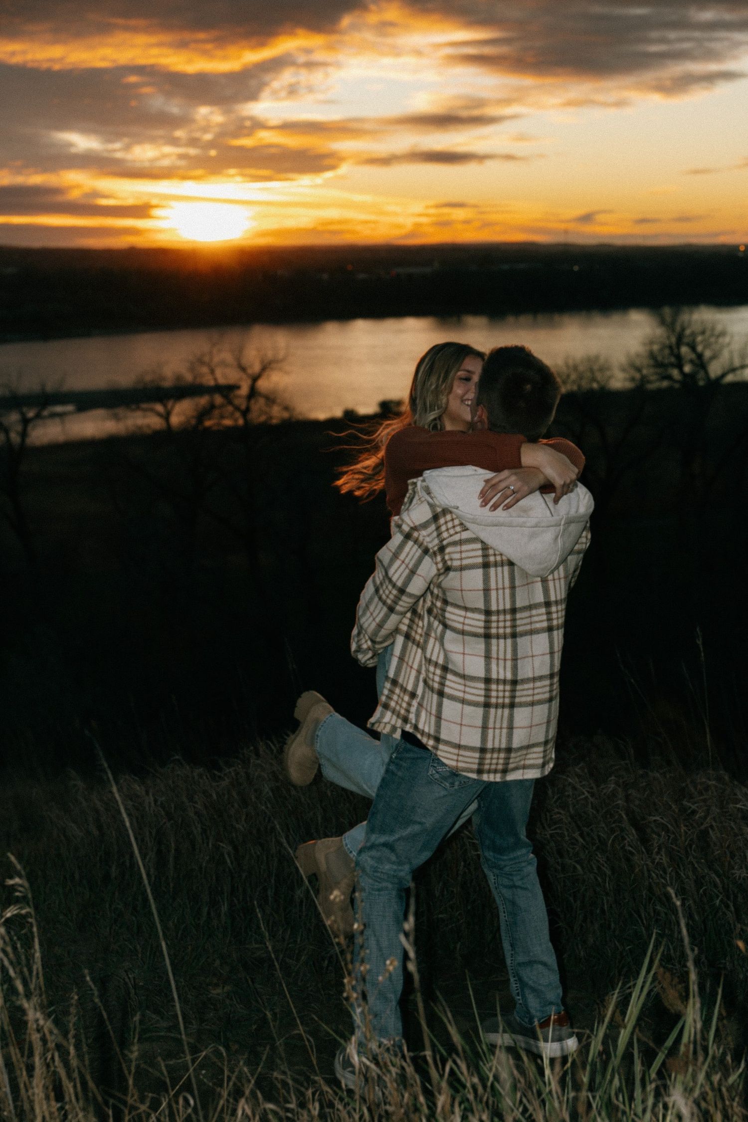 Silhouettes embrace during a dramatic sunset by a lake with tall grass.