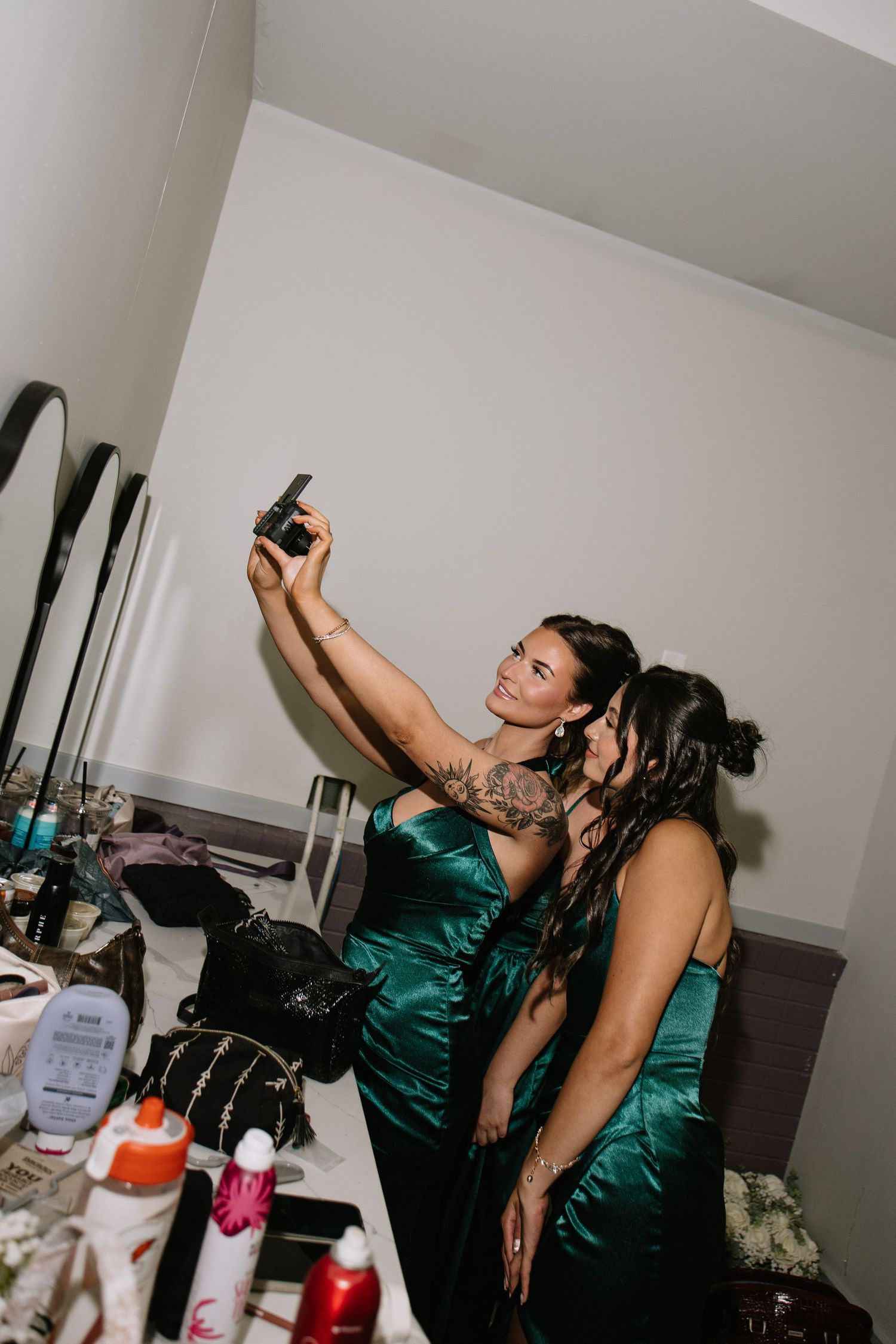 Two women in emerald green satin gowns taking a selfie at a vanity mirror during wedding preparations.