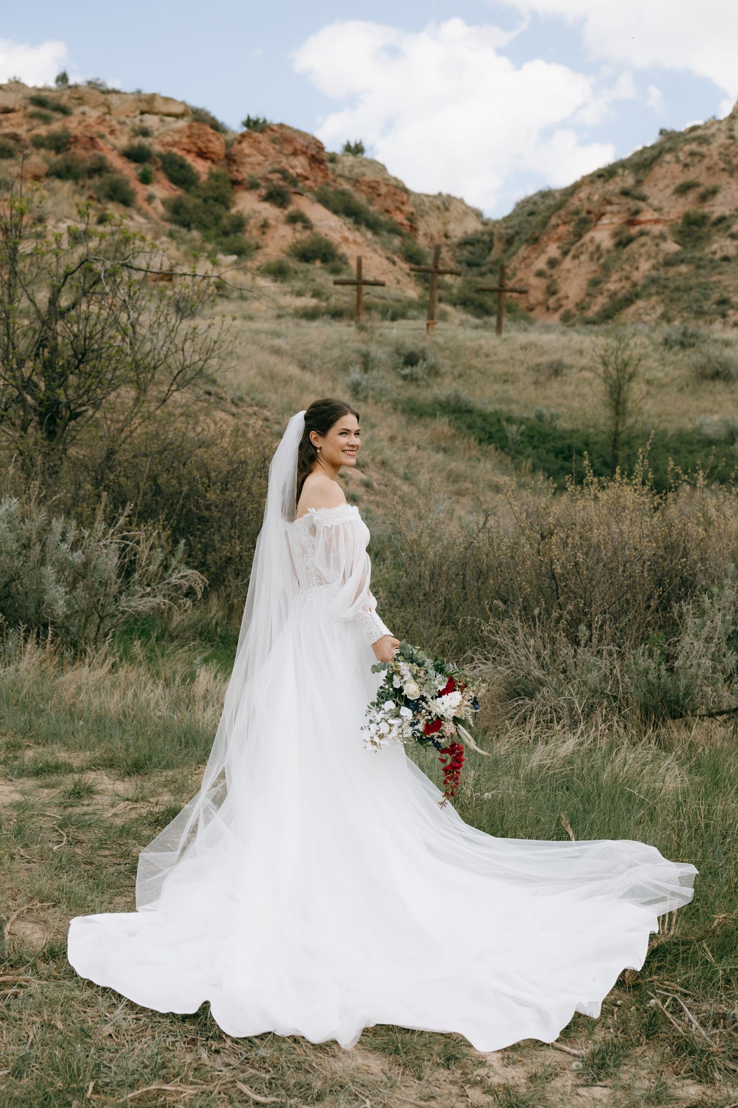 Elegant bride in a white gown and veil holds a bouquet in a scenic outdoor landscape with rocky hills.