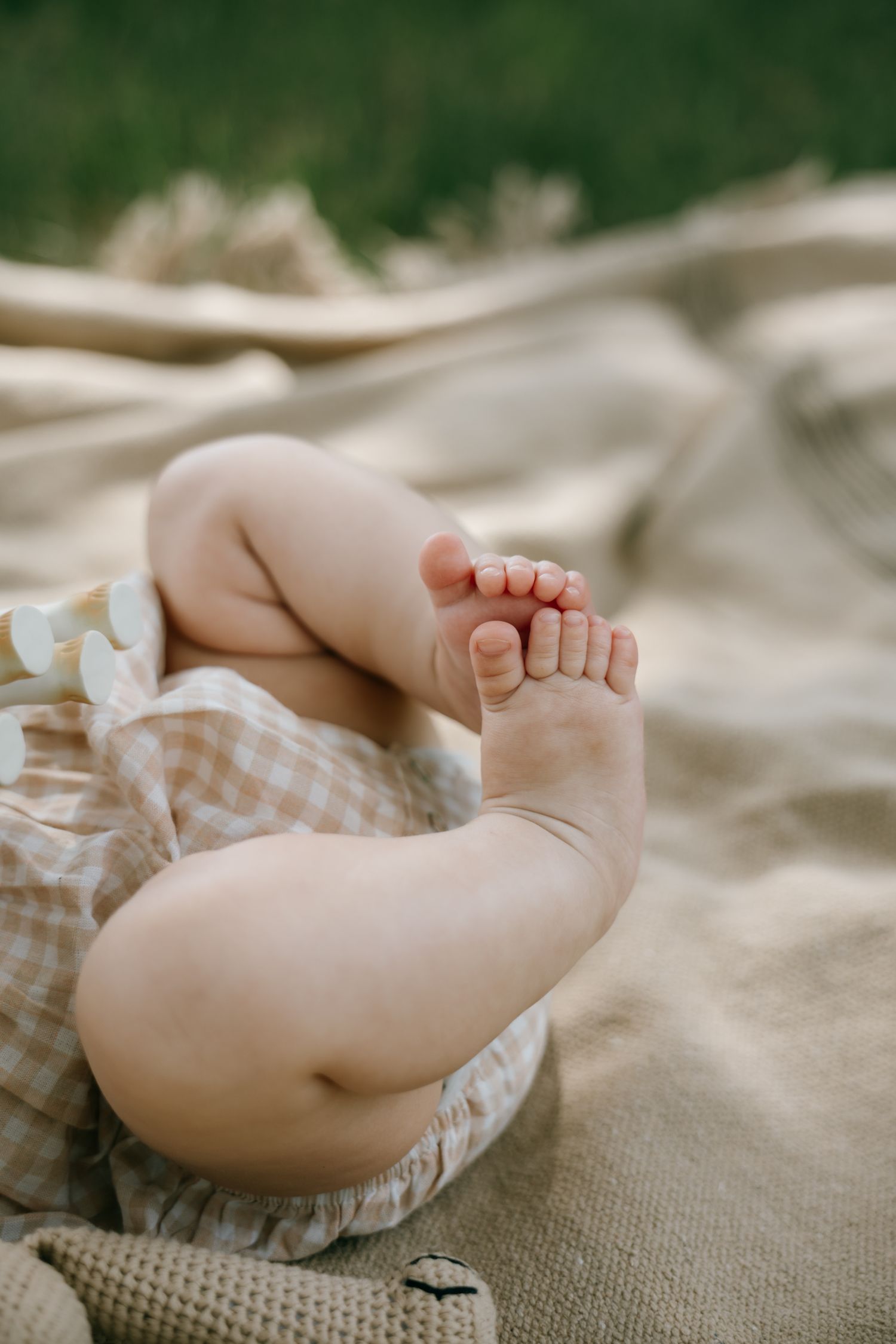 Close-up of chubby baby feet and legs on a beige knit blanket outdoors, wearing a gingham outfit.
