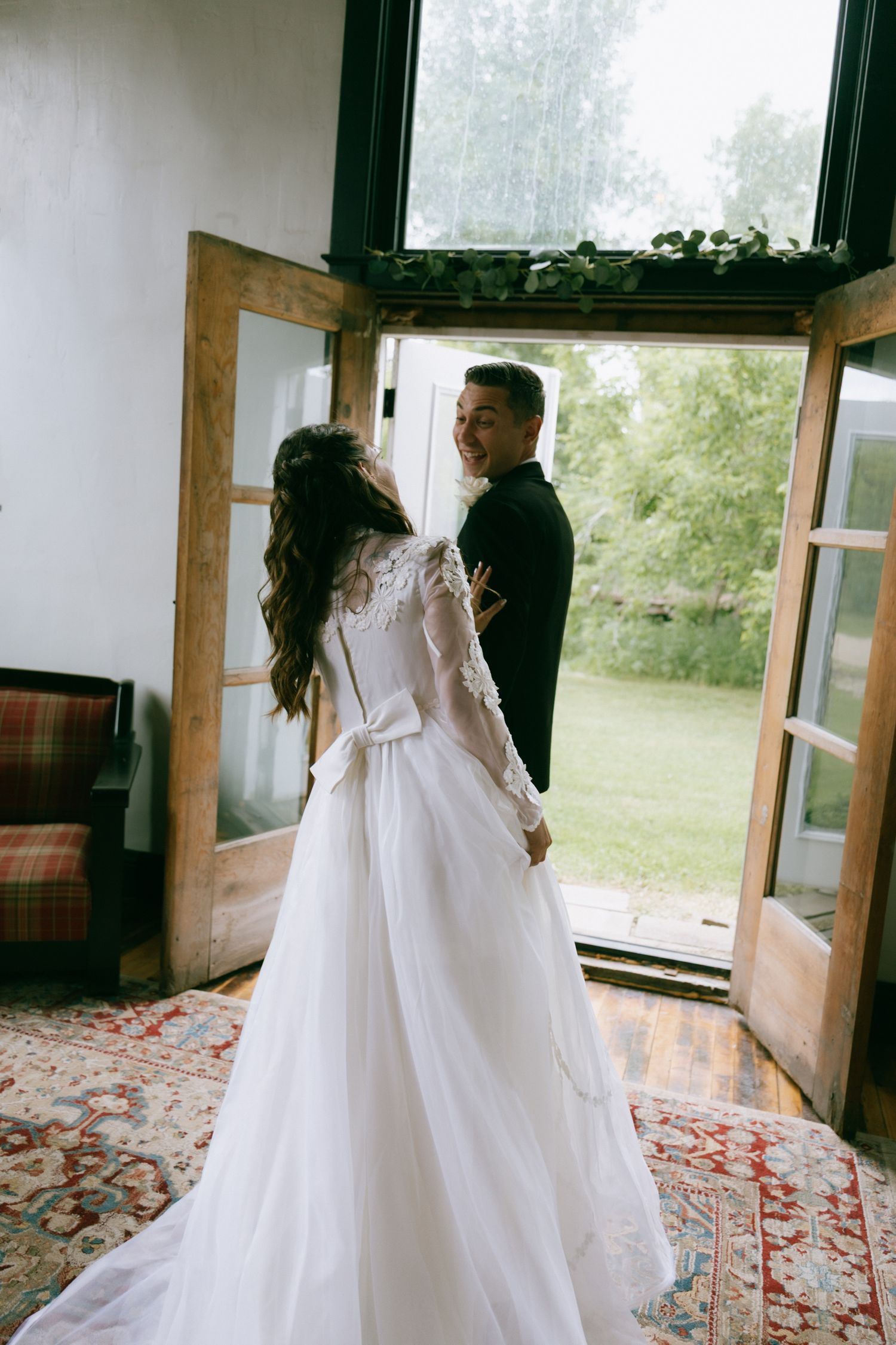 Bride in lace wedding gown and groom in black suit stand at open wooden doors overlooking a lush green garden.
