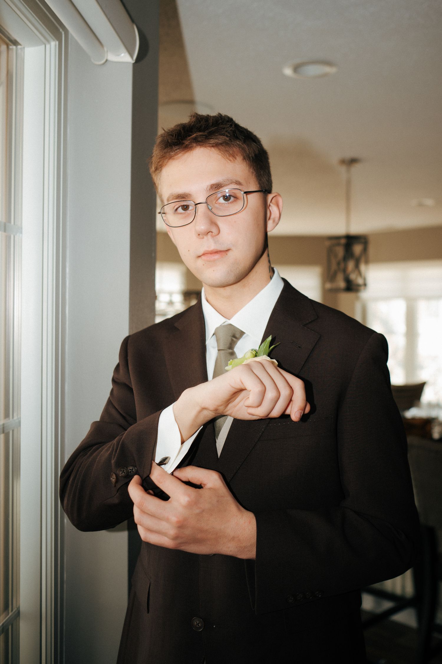 Young man in dark suit adjusting boutonniere, wearing glasses and olive tie, standing by window in formal attire.
