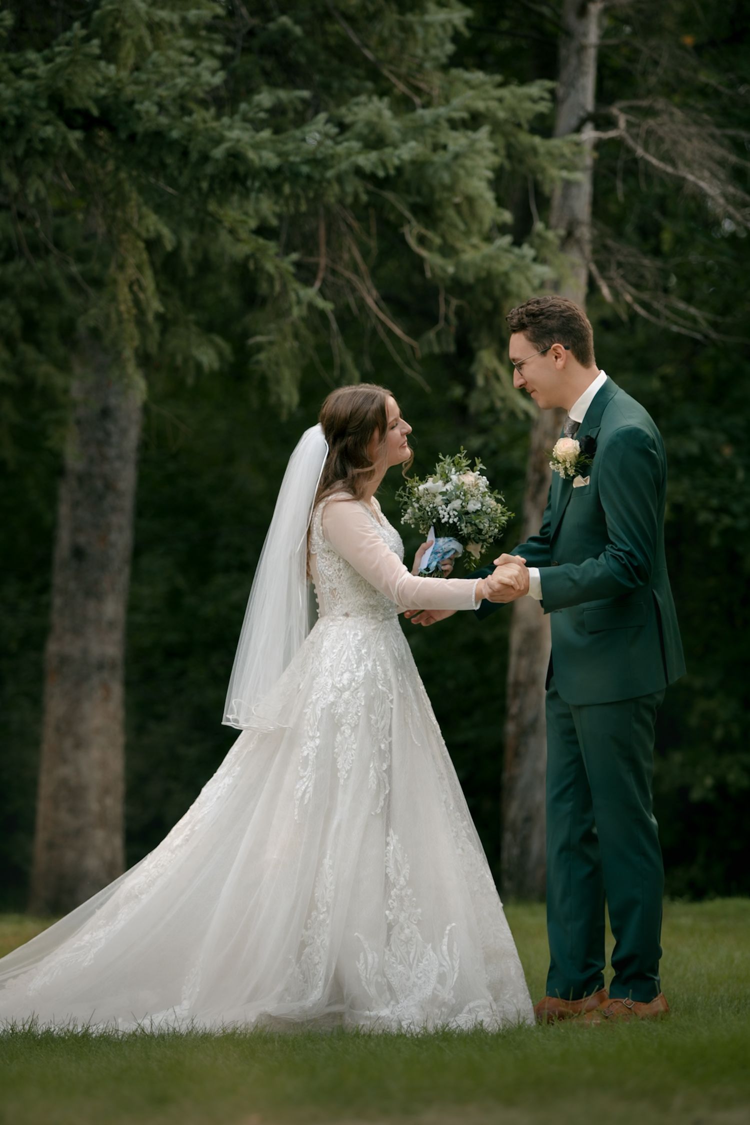 Wedding couple exchanging flowers during outdoor ceremony surrounded by tall trees.