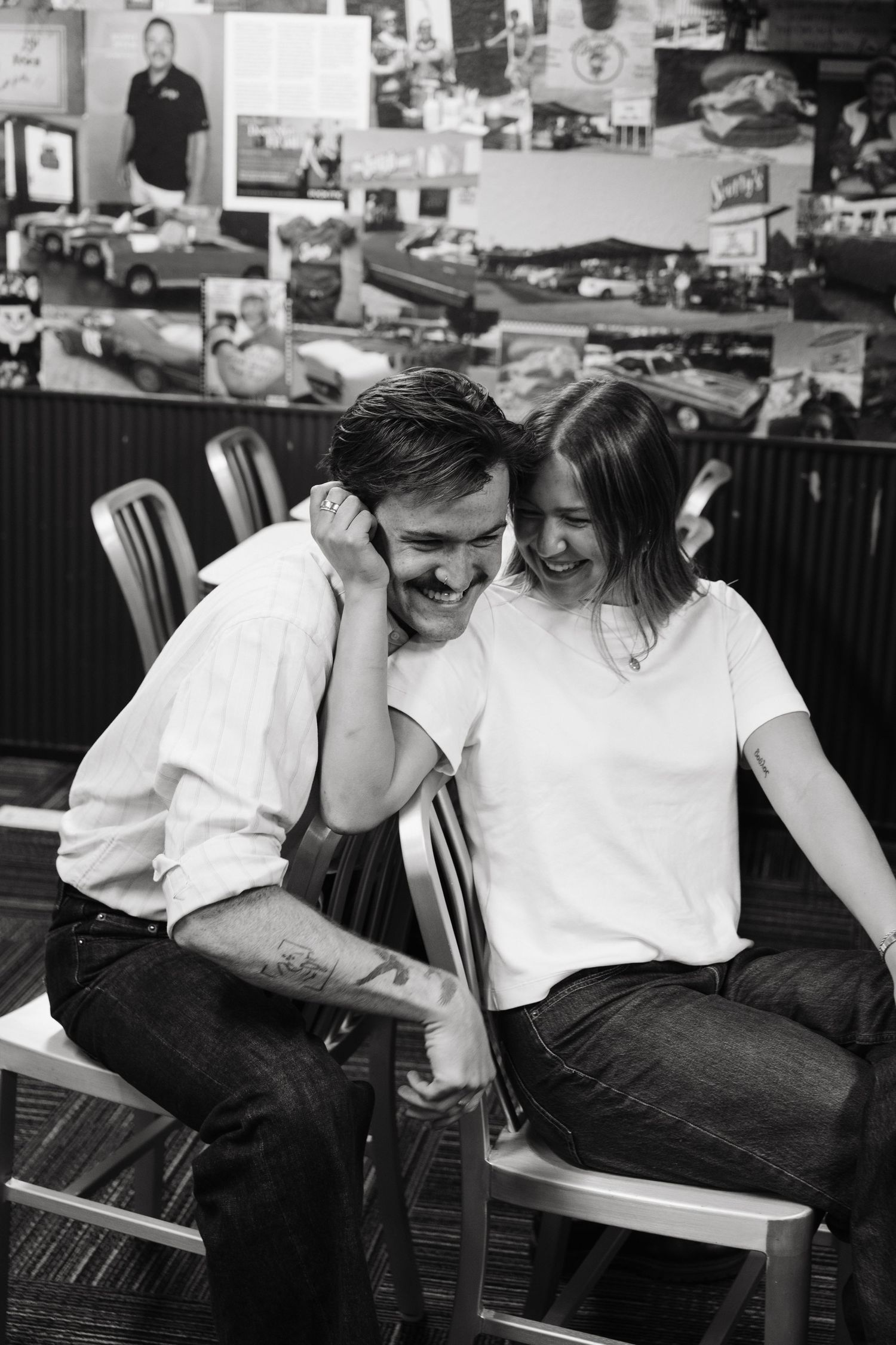 Laughing couple sitting close together in a diner, black and white photo with memorabilia wall behind them.