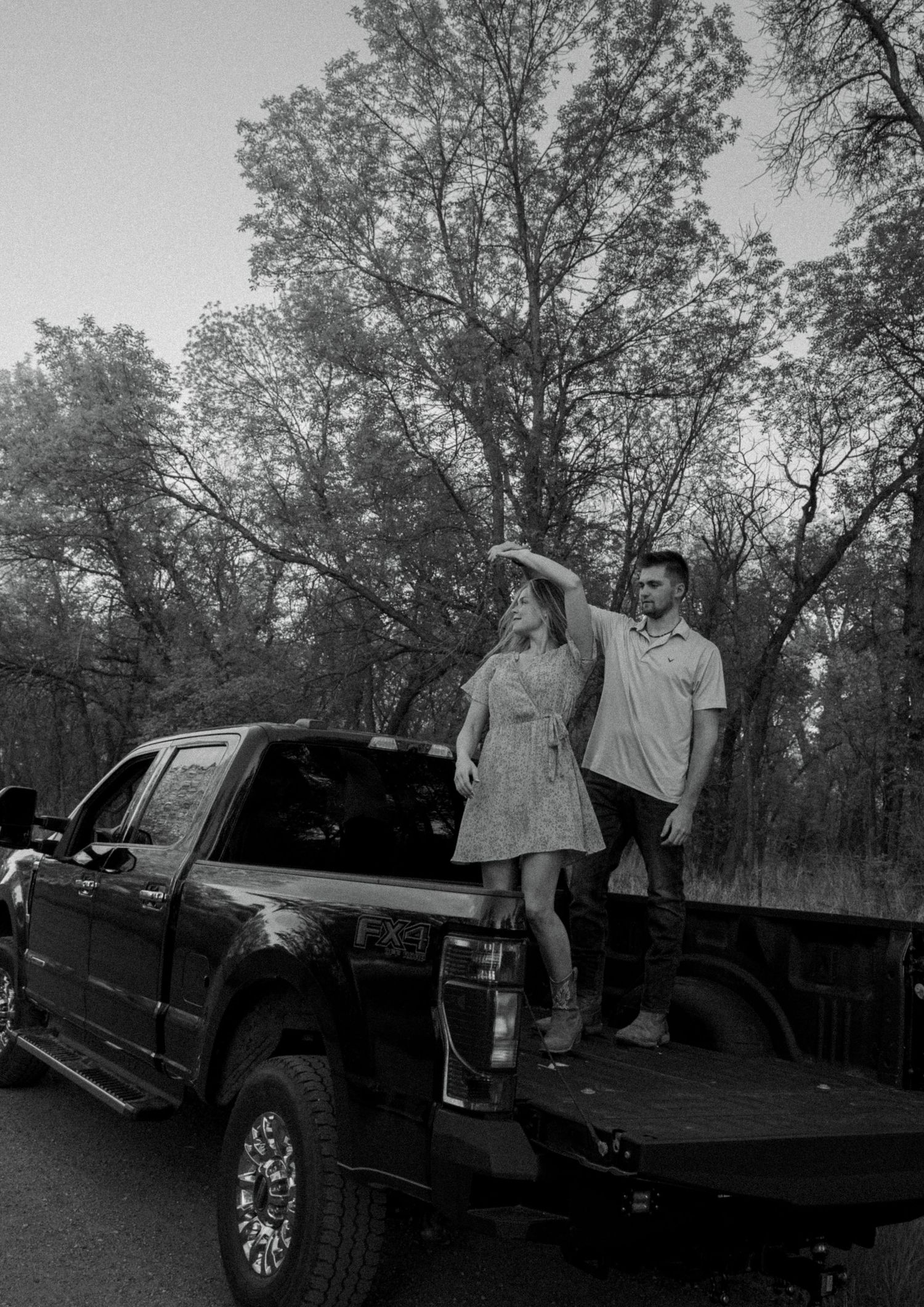 Couple poses together near black pickup truck in outdoor wooded setting.