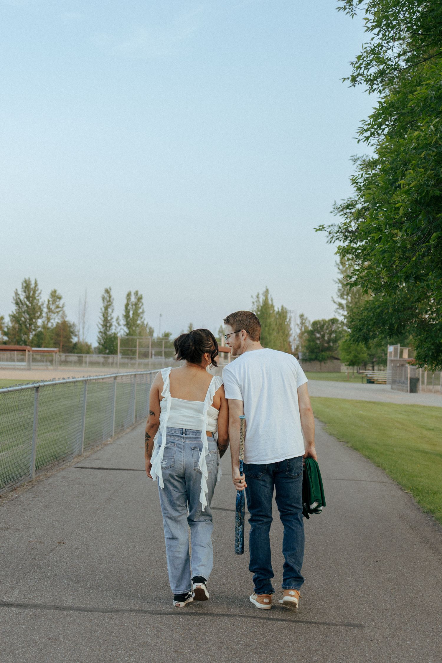 Two people walk hand in hand down a path beside a chain link fence.