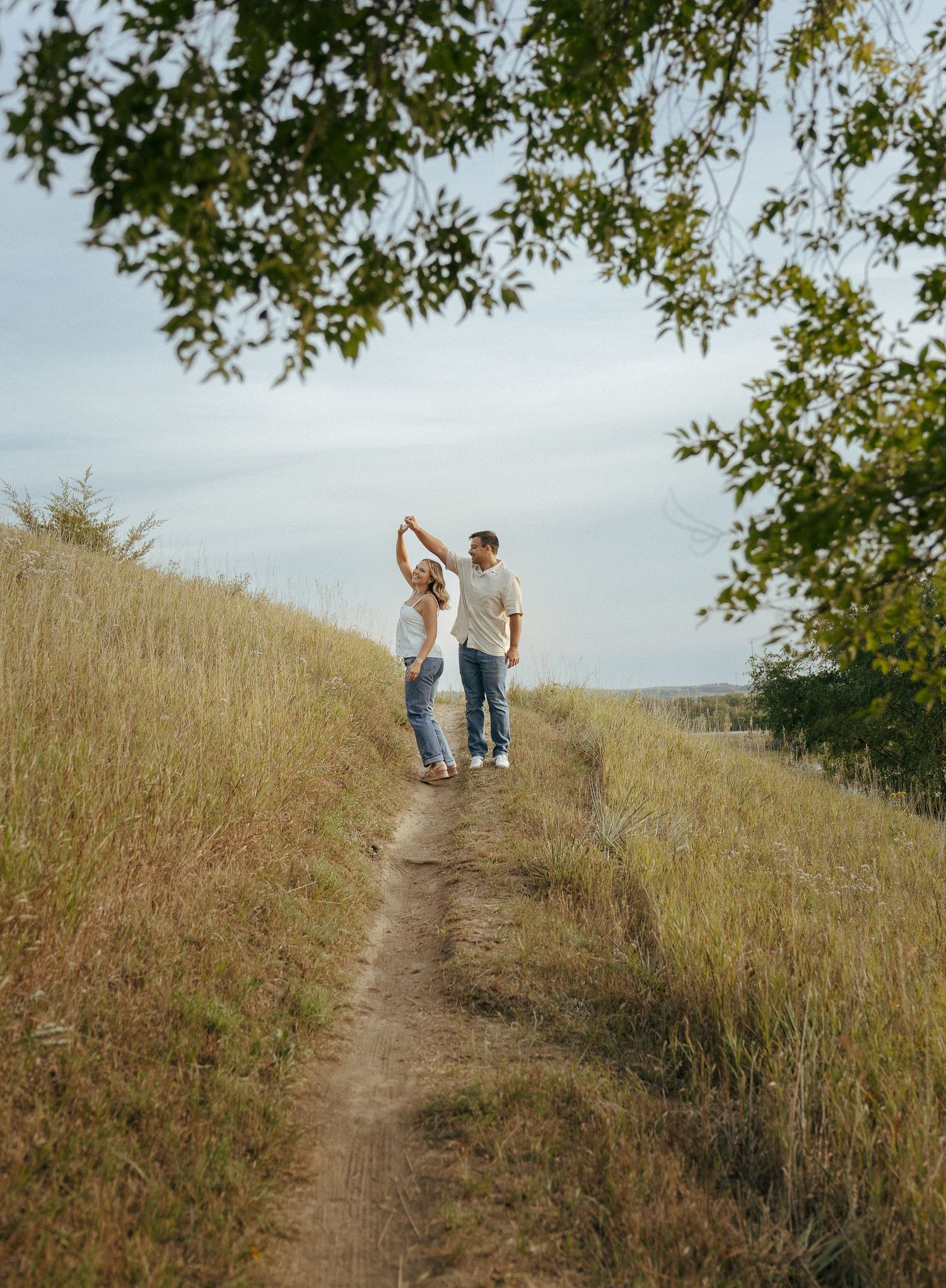 Two people stand together on a winding dirt path through tall grass and trees.