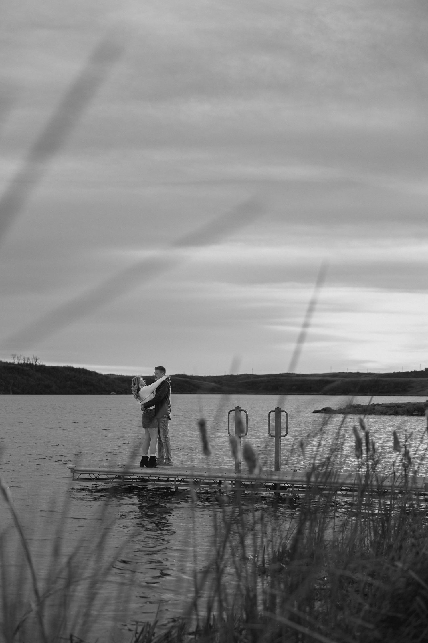 A black and white photograph captures two people on a dock over calm waters.