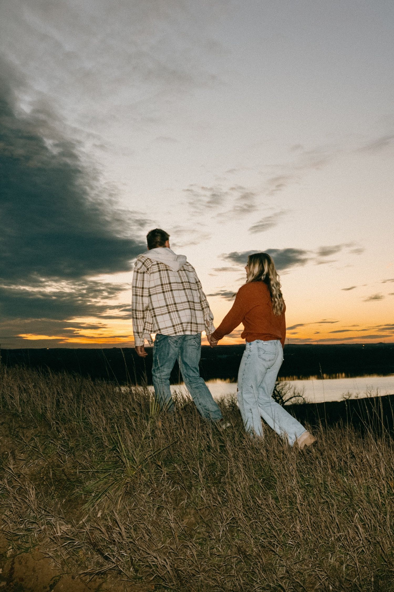 Couple walking hand in hand through field at dusk with water view in background.