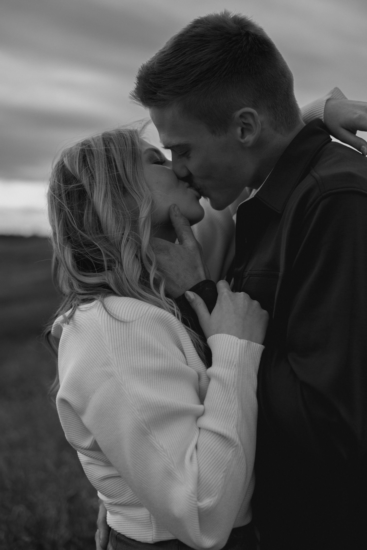 A romantic black and white portrait of a couple sharing an intimate kiss outdoors at sunset.