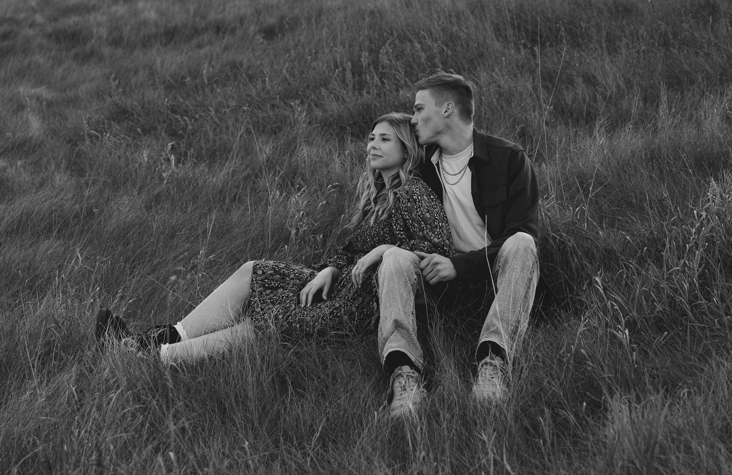 Couple lying together on grassy hillside during romantic outdoor photoshoot.