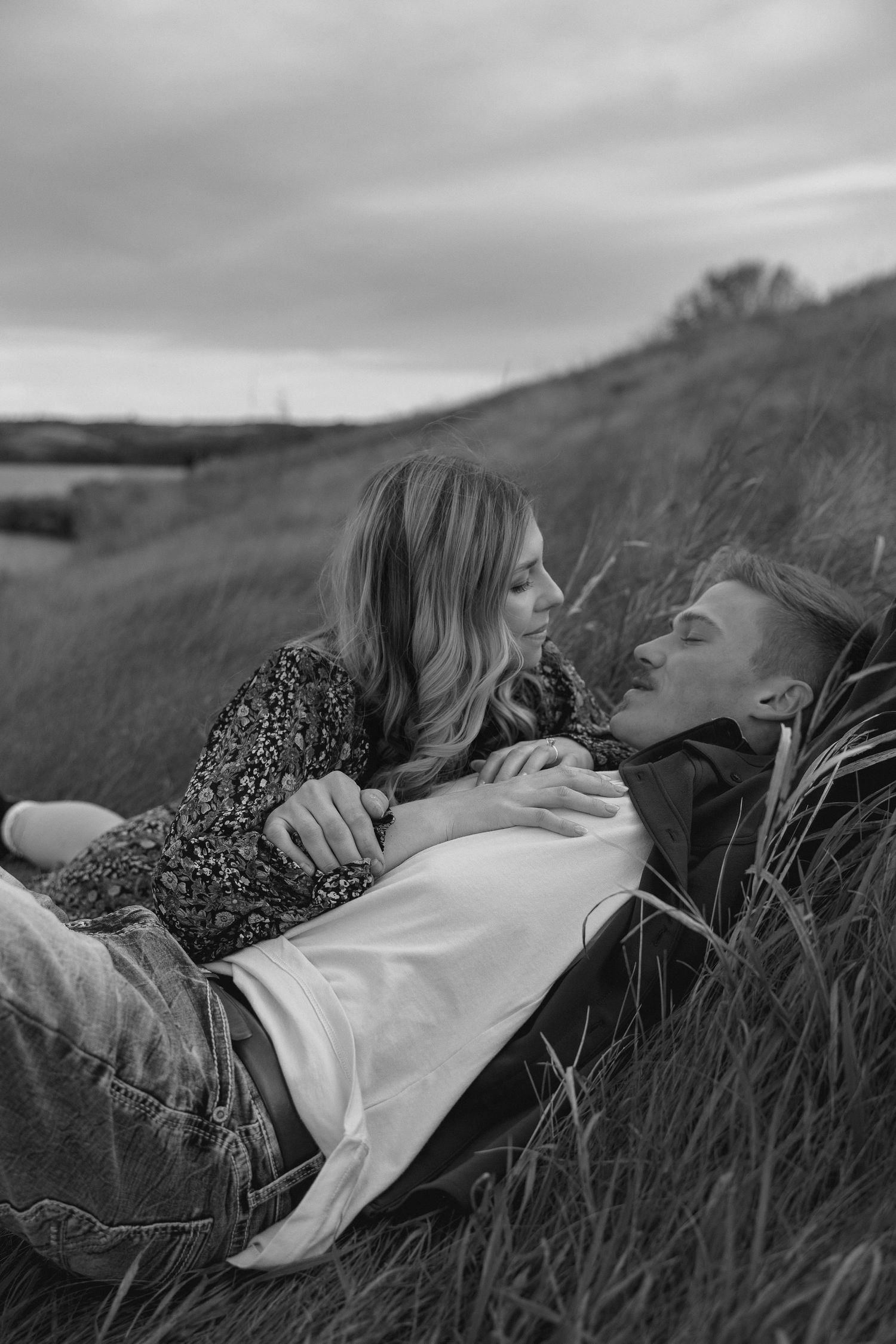 Couple lying together in tall grass field on cloudy day, shot in black and white.