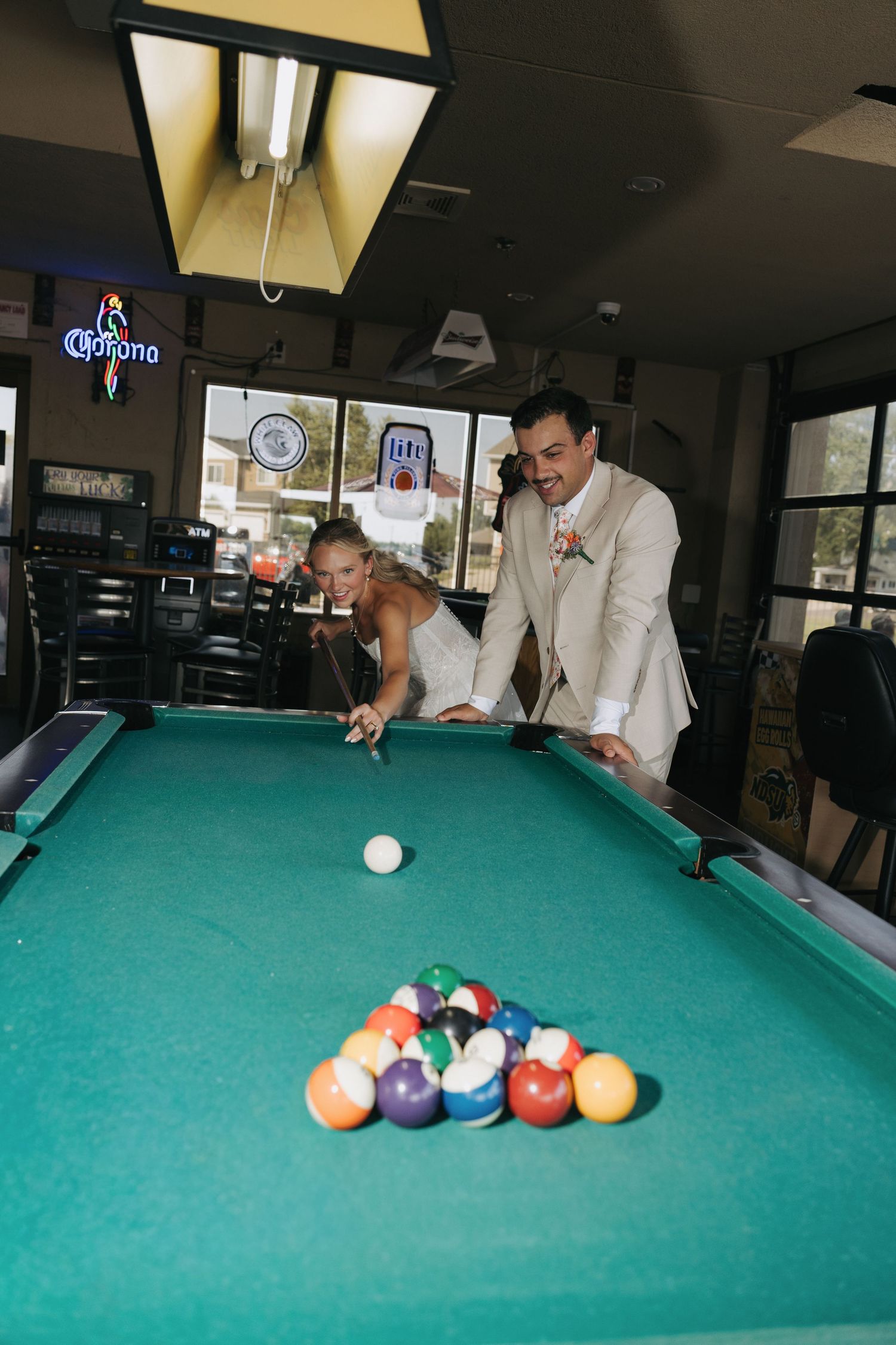 Newlywed couple in wedding attire playing pool billiards at a bar with Corona neon sign in background.