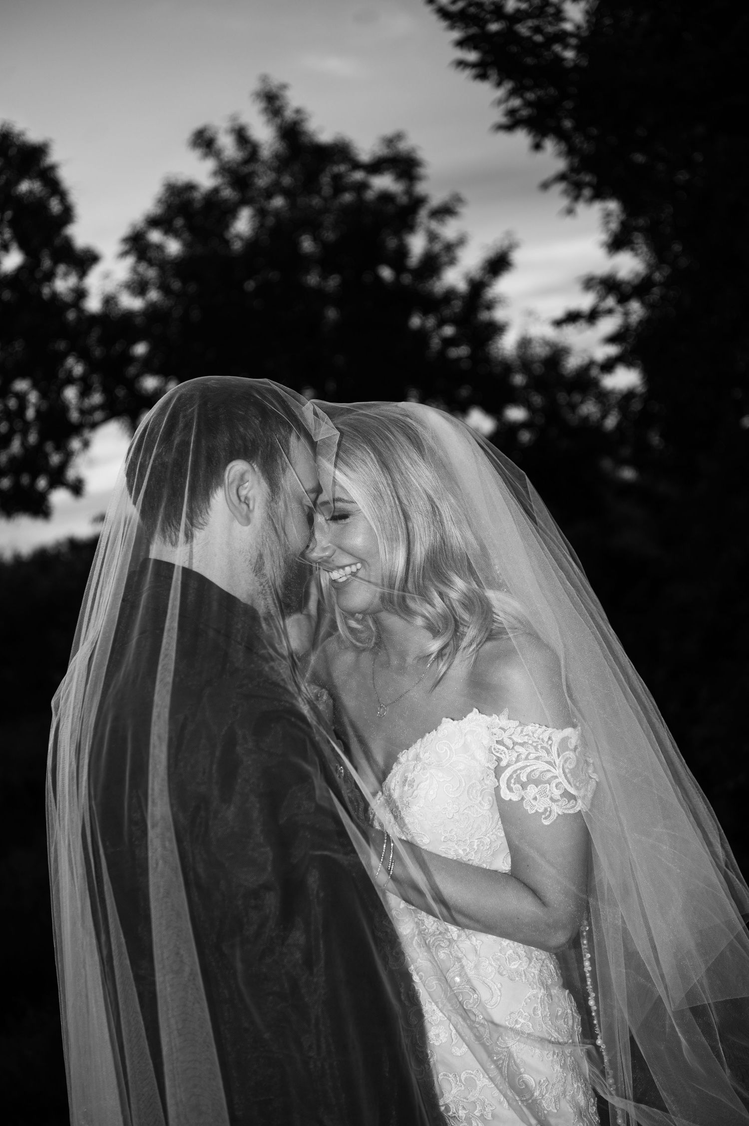 Black and white photo of a bride and groom sharing a joyful moment under her veil outdoors.