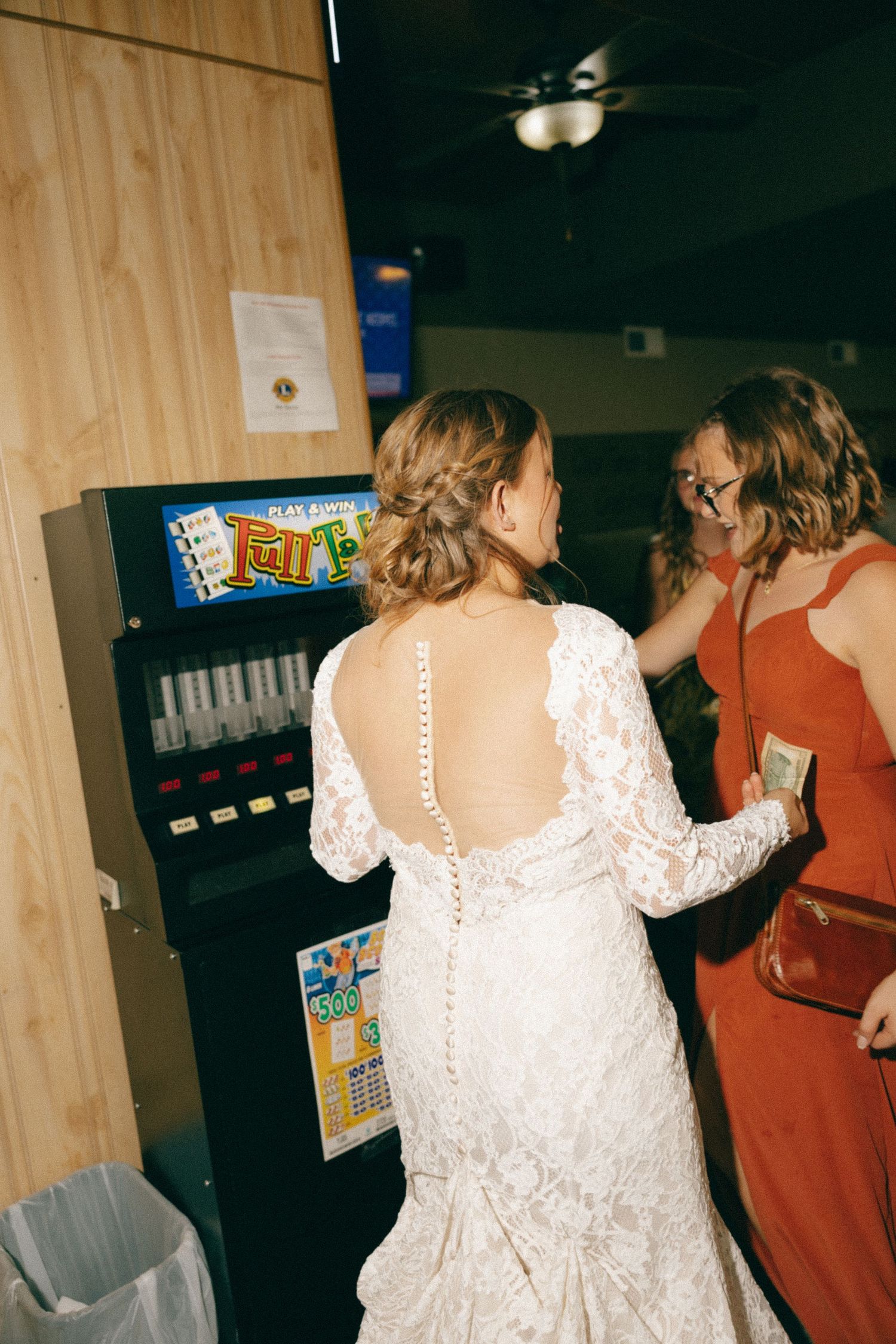 Bride in lace wedding dress with button back at lottery machine with guest in red dress.
