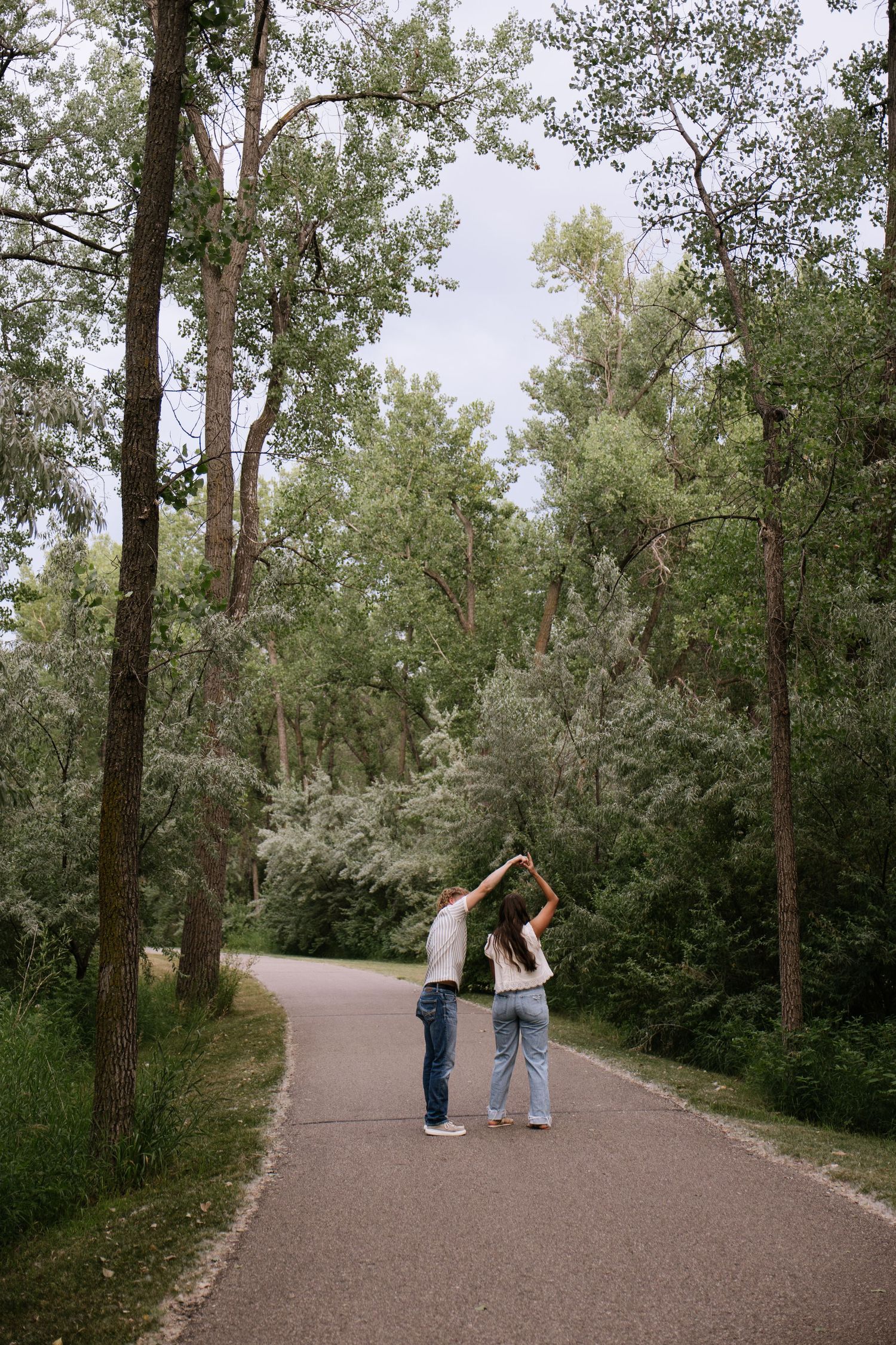Couple dancing together on a tree-lined park path, holding hands raised, wearing casual jeans and t-shirts on a cloudy day.