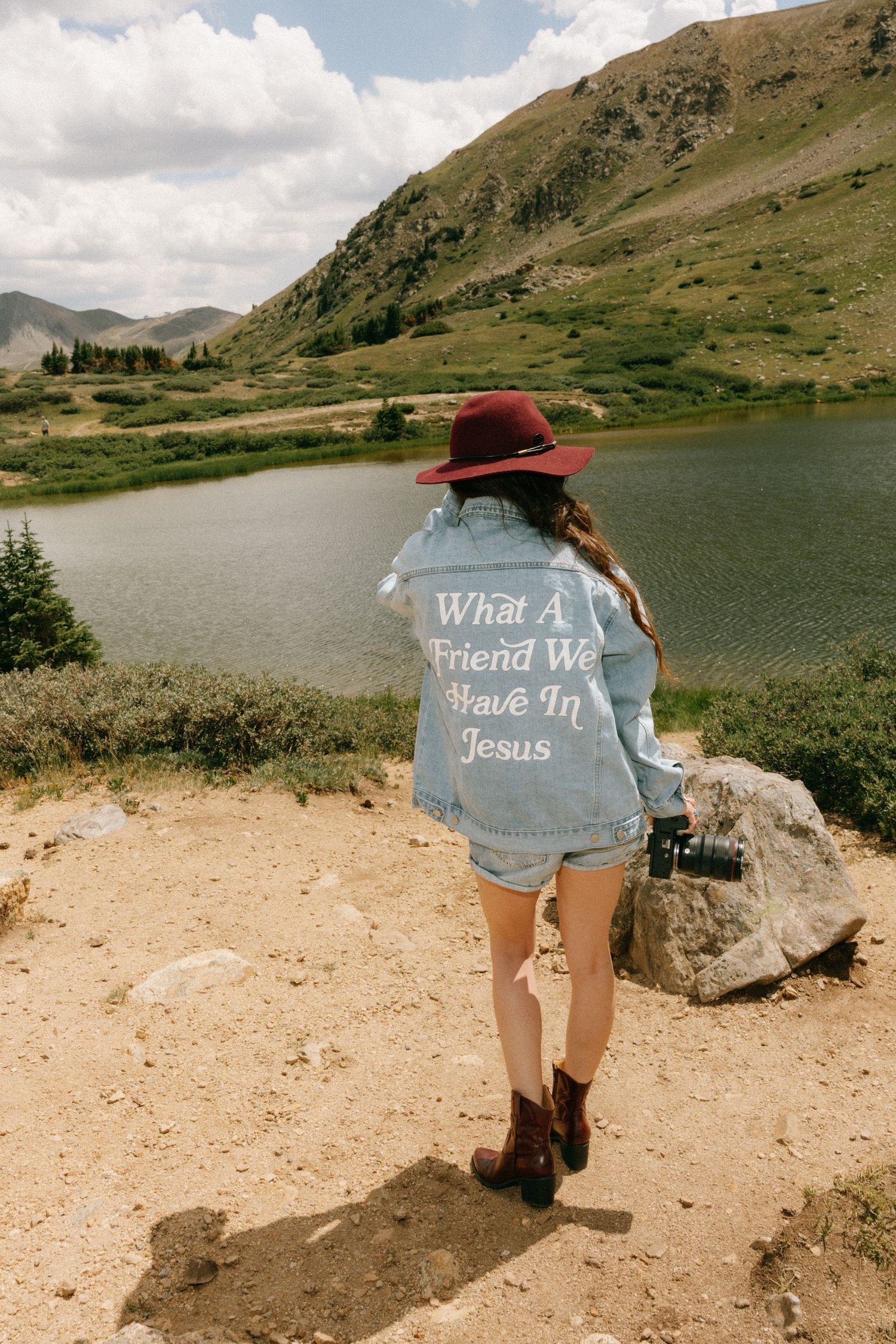 Woman in denim jacket reading What A Friend We Have In Jesus stands by mountain lake wearing red hat and boots.