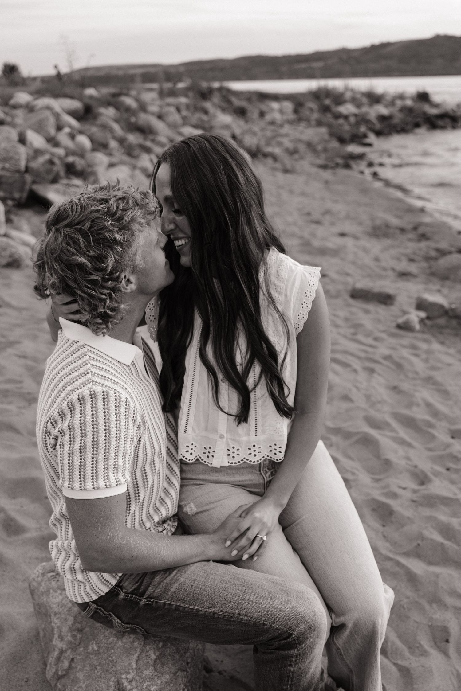 Black and white photo of a couple sharing a romantic moment on a rocky beach, smiling and leaning in close.