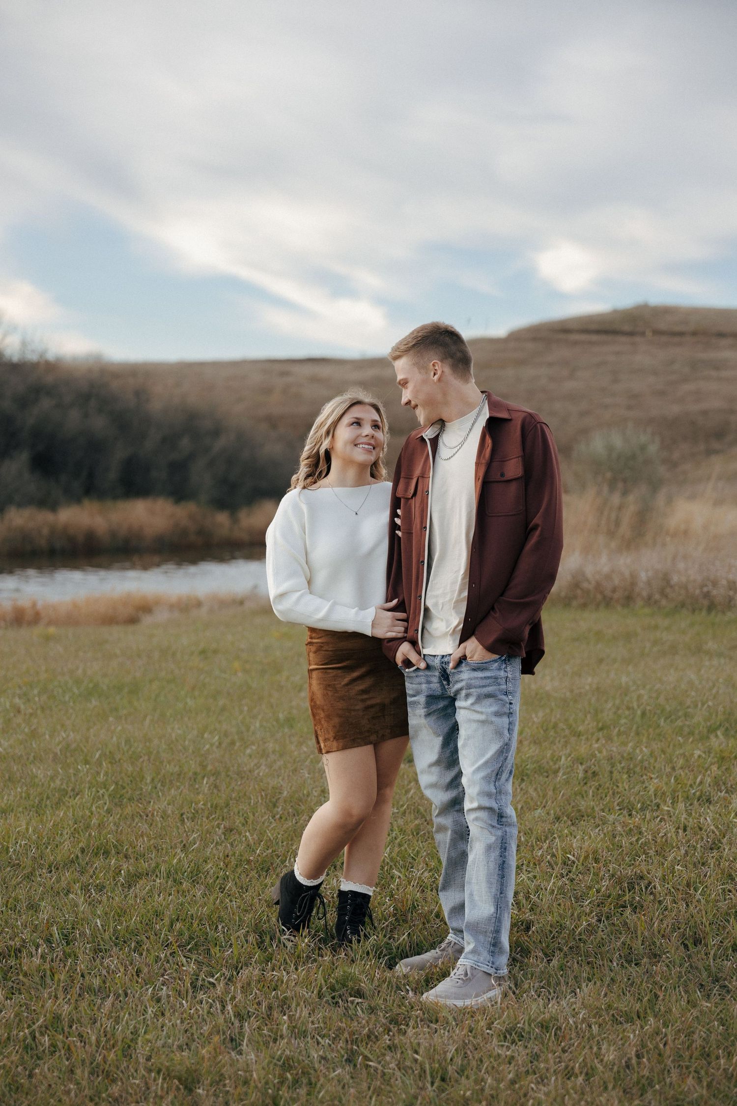 A couple in casual fall attire stands together in a grassy field with mountains in the background.