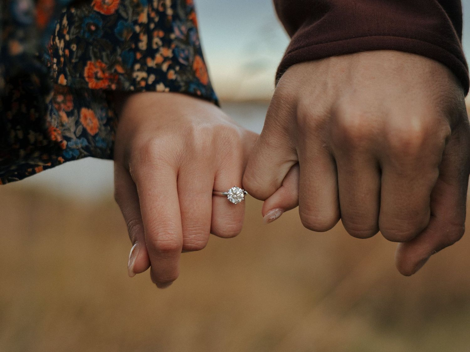 Close-up of hands with engagement ring showing intertwined fingers against floral fabric.
