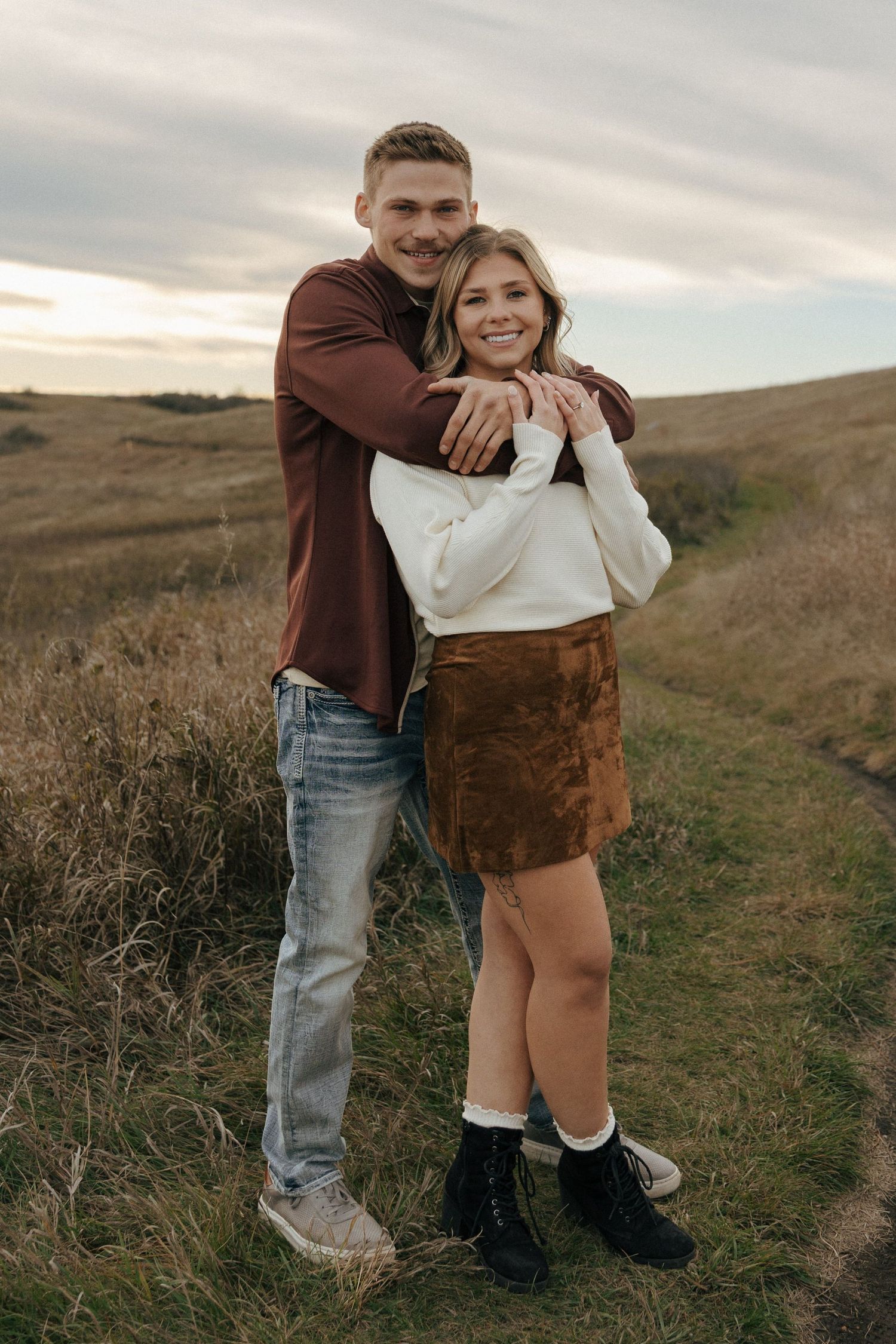 Playful couple embracing on a grassy hillside trail.