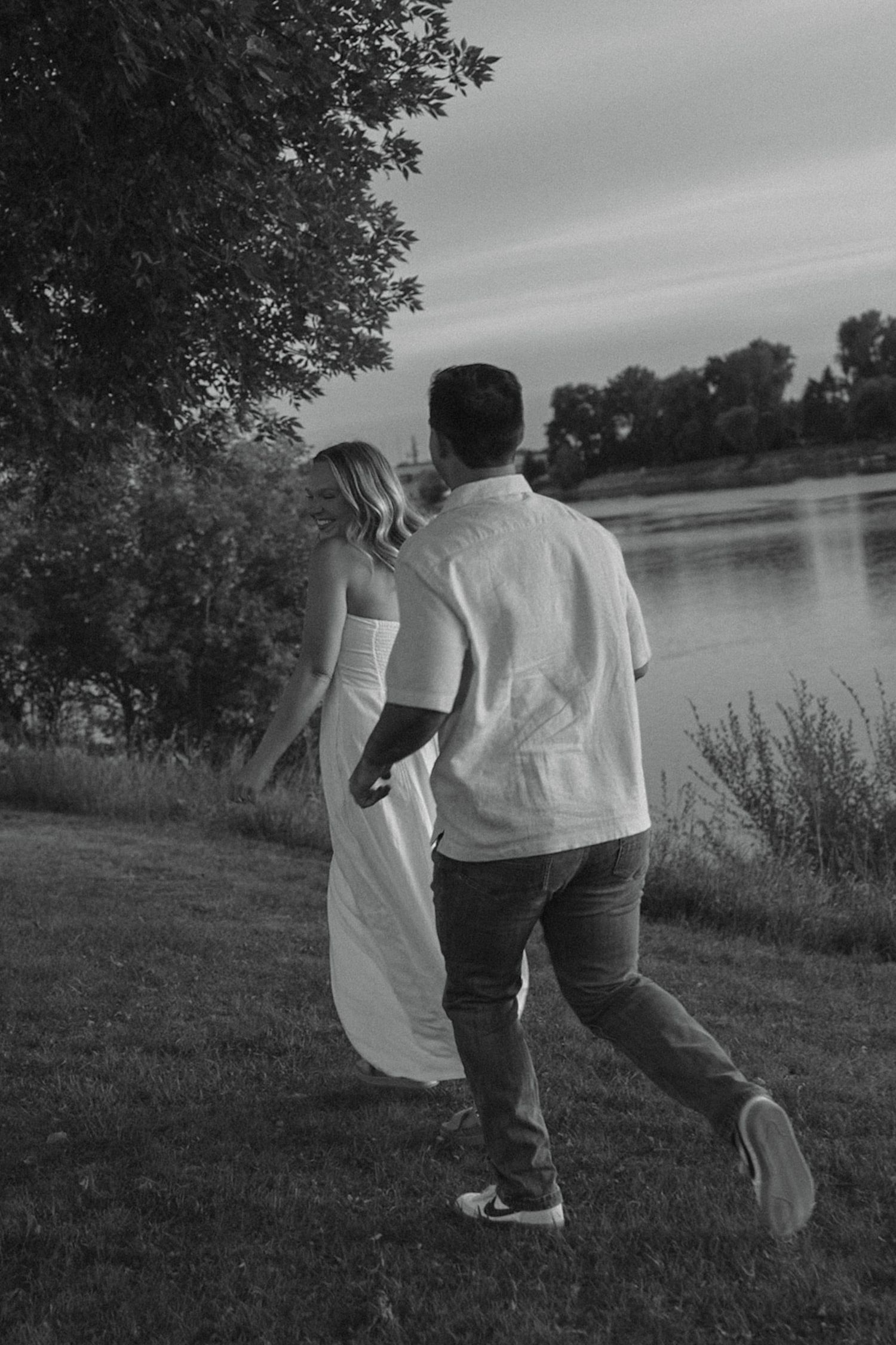A couple walks together along a lakeside path during sunset, holding hands in casual white outfits.