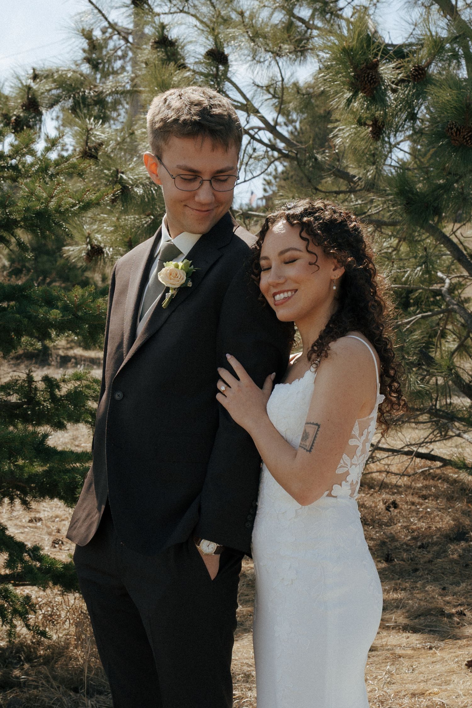 Bride in white lace gown and groom in black suit with boutonniere pose outdoors near pine trees on wedding day.