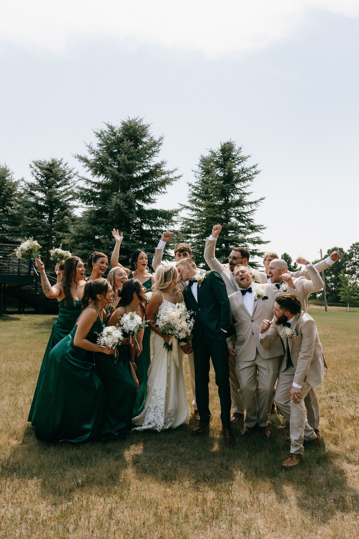 Joyful wedding party celebrating outdoors in green bridesmaid dresses and tan suits with evergreen trees behind them.