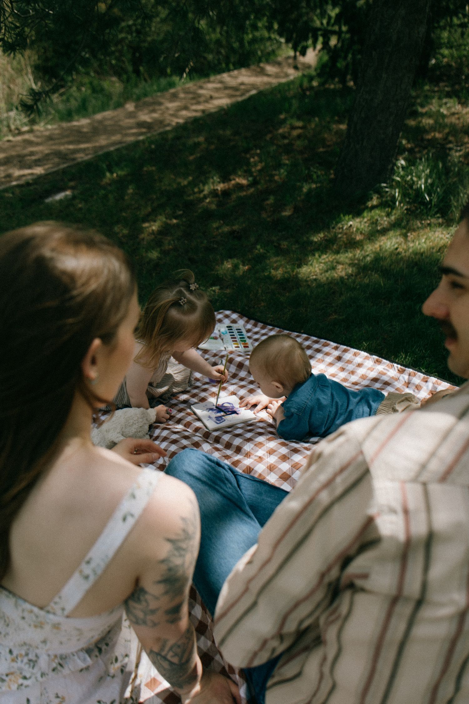 Family enjoying a sunny outdoor picnic on a checkered blanket in the shade of trees with children playing.