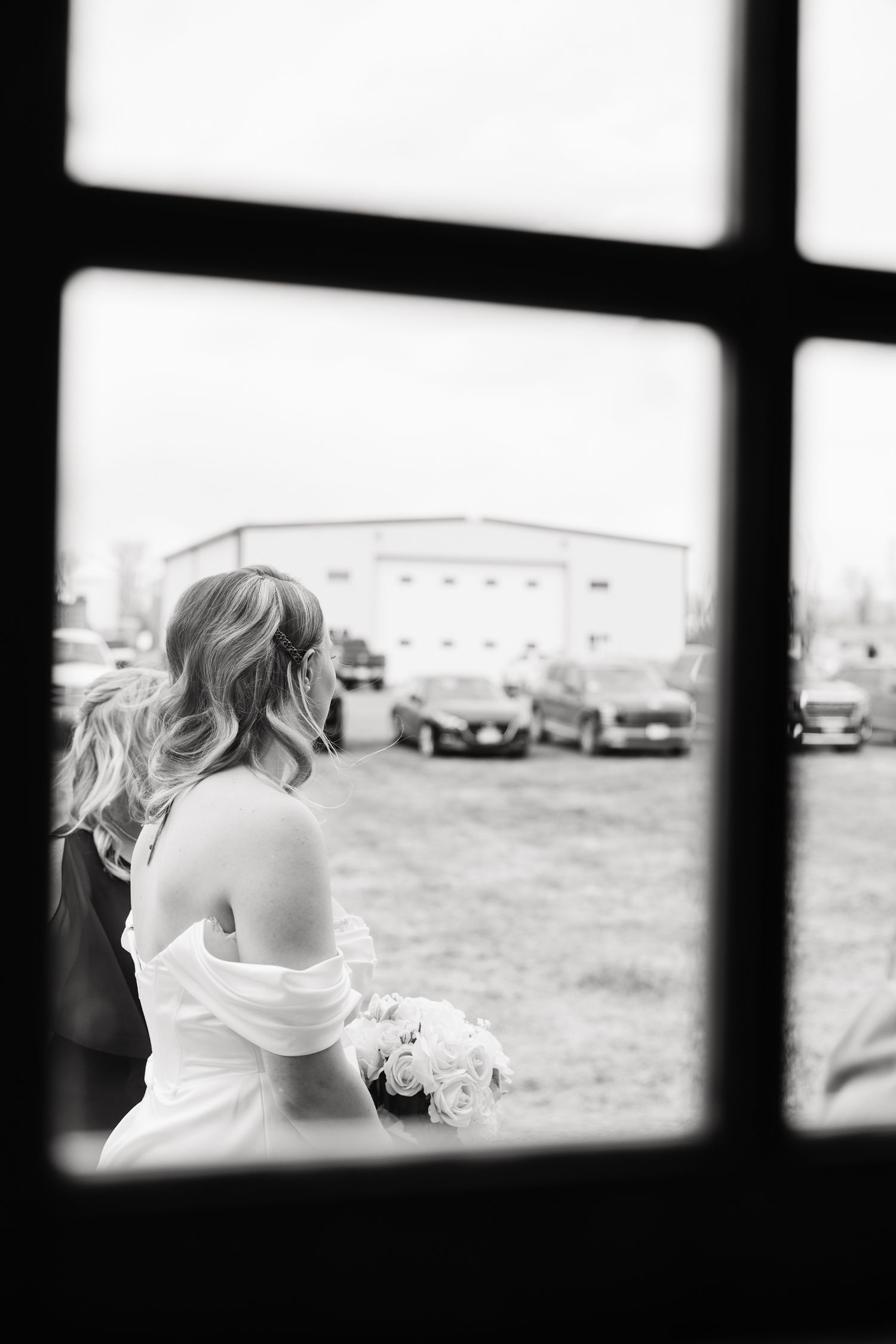 Black and white photo of a bride in an off-shoulder gown holding a bouquet, viewed through a window.