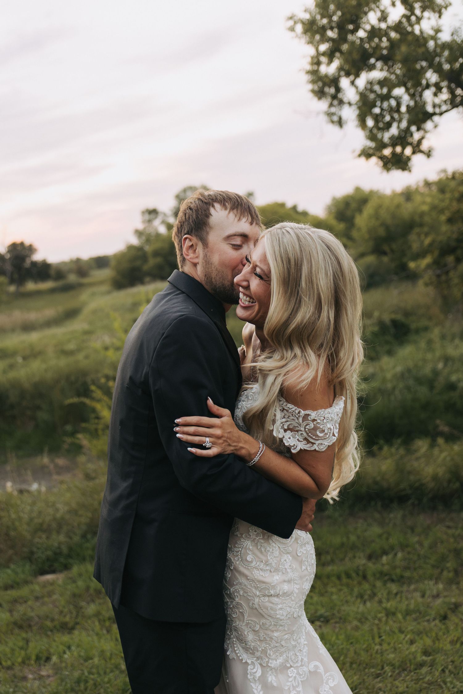Bride and groom share a romantic kiss outdoors at sunset, surrounded by lush green fields and trees.