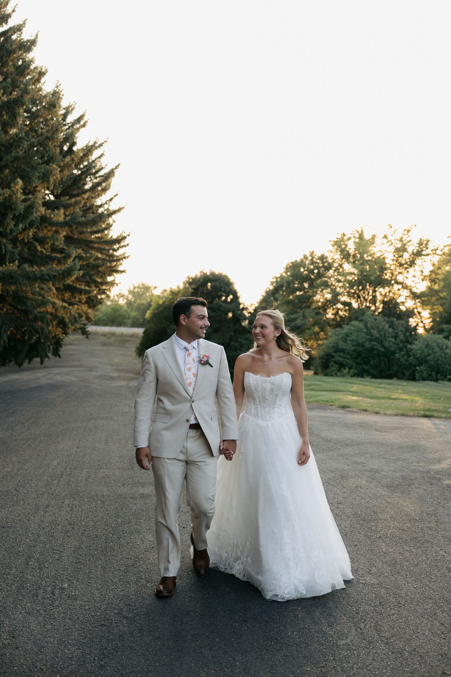 A smiling bride and groom walk hand in hand down a tree-lined path in golden hour light on their wedding day.
