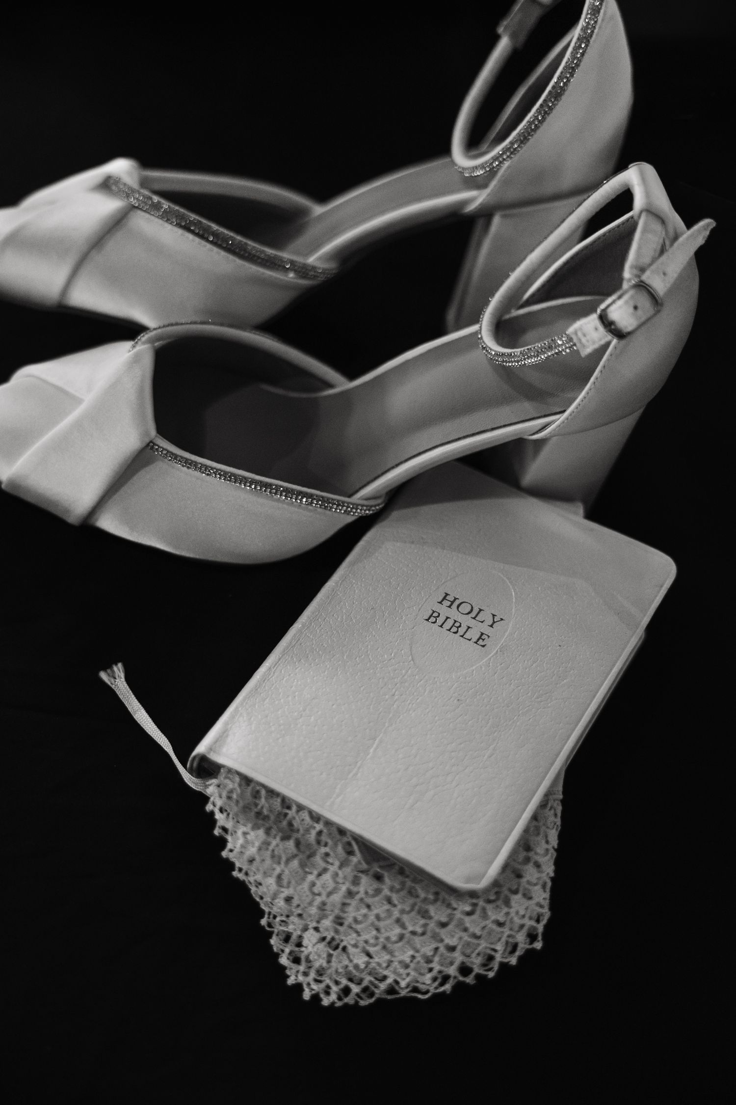 Black and white photo of bridal heels, Holy Bible, and lace handkerchief on dark background.