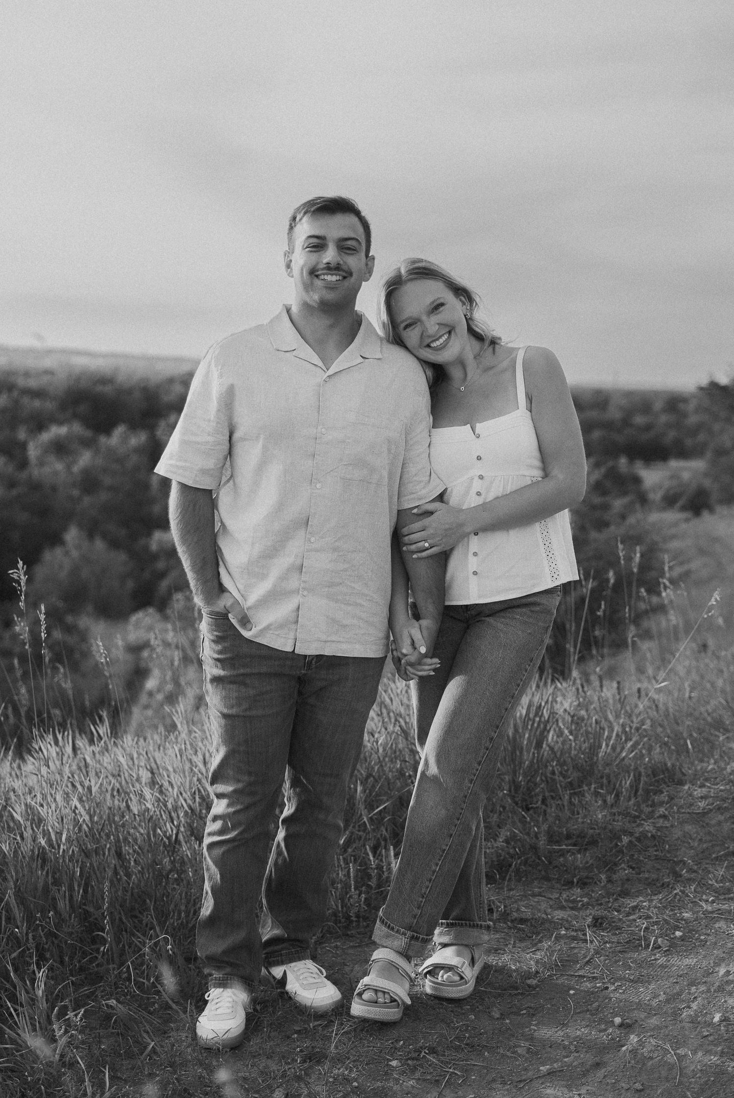 A couple wearing white tops and jeans poses together on a dirt path surrounded by tall grass.