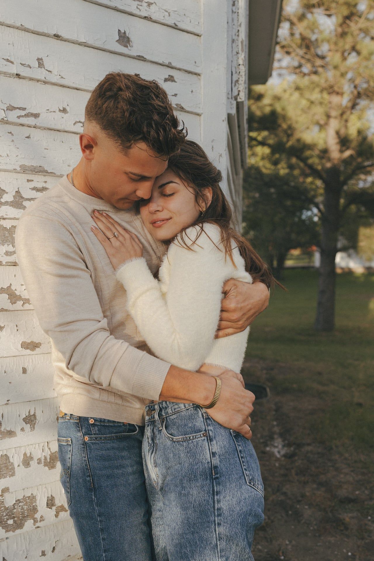 A romantic couple in casual attire embraces tenderly against a white wall in an outdoor autumn setting for an engagement session.
